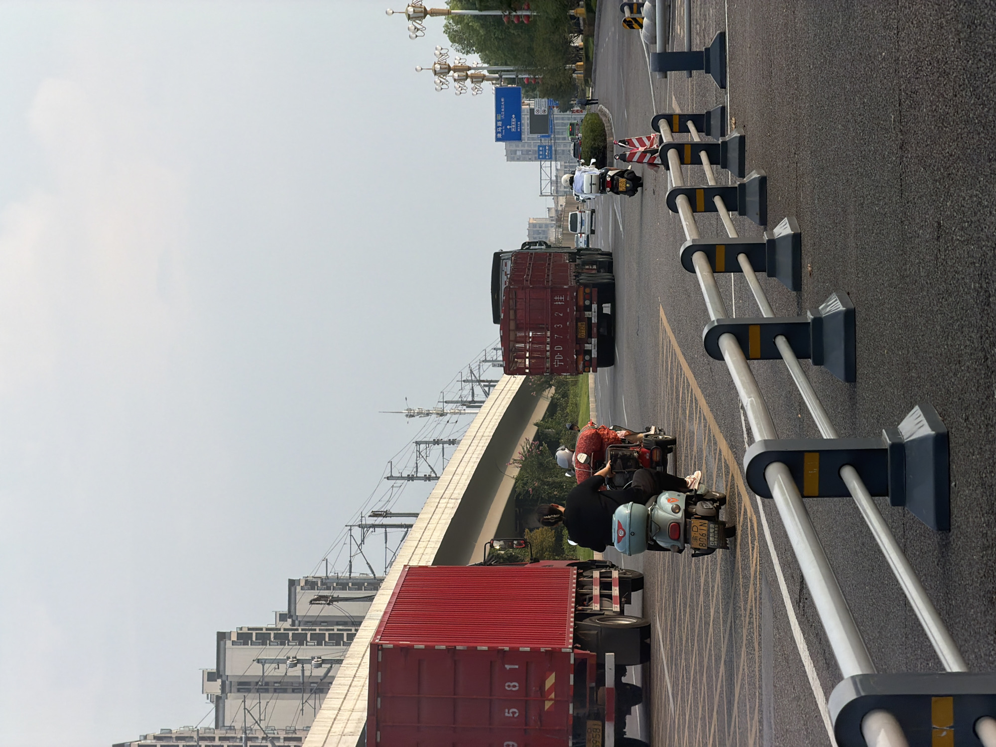 In Jiangjin, China, a street scene unfolds under a pale, overcast sky. A multi-lane road features a variety of vehicles. A large red truck, numbered 9581, dominates the left side of the frame, its trailer emblazoned with more numbers. Ahead of it, a red trailer truck, identified as 宁D.D 732 挂, travels in the same direction.
In the foreground, a light blue scooter is ridden by a person wearing a black top and dark pants. Behind them, a three-wheeled vehicle, loaded with produce, carries two passengers, one wearing a white helmet. The road is marked with yellow and white lines, and a modern barrier system with dark bases and silver rails separates the lanes.
In the background, the elevated tracks of a railway line cut across the image, supported by concrete structures. Apartment buildings with a uniform architectural style line the horizon on the left. On the right side of the road, decorative streetlights with multiple lamps are spaced along the sidewalk, and a blue directional sign points towards various locations within Jiangjin, including 滨江大道. Trees and vegetation border the road. The overall atmosphere suggests a typical urban environment in motion.