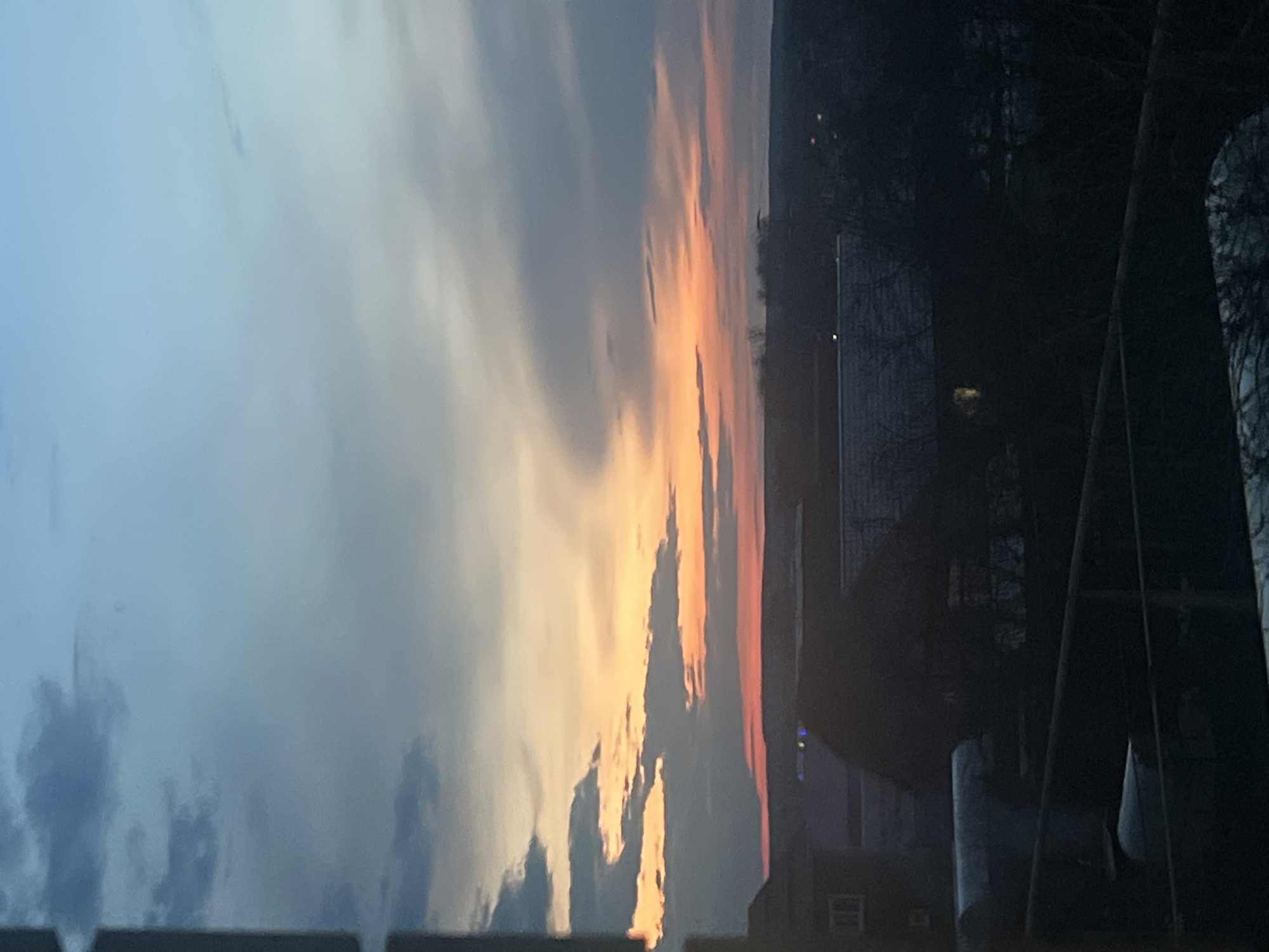The image displays a serene sunset or sunrise over a landscape, with the horizon line spanning horizontally across the mid-section. The upper portion of the frame is dominated by a dramatic sky, showcasing a gradient of colors from soft blues and grays in the upper left to brilliant oranges, yellows, and pinks along the horizon, suggesting the low angle of the sun. Various clouds are present, reflecting the vibrant light.
Below the horizon, the foreground is rendered in deep silhouette due to the backlighting. Dark, indistinct shapes of bare trees are visible on both the left and right sides, suggesting a wooded area. In the mid-ground, faint outlines of structures, possibly buildings or houses, are discernible as dark masses against the sky's glow. On the far right, a dark, vertical element, potentially a utility pole or part of a structure, stands out. The scene evokes a tranquil atmosphere at either dawn or dusk, in what appears to be a rural or suburban setting in Votkinsk, Russia. There are no people or discernible text in the image.