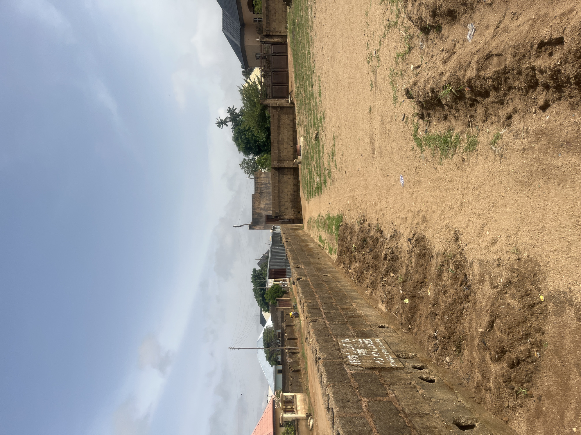 The image shows a street in Jos, Nigeria, with a partially visible compound wall on the right. The sky is visible on the left, with a mix of light blue and cloud cover. The ground is sandy and shows evidence of recent digging or disturbance. Buildings of various sizes and architectural styles are visible beyond the wall and along the street. A utility pole with wires is on the left side of the image. A sign is visible on the wall, although the text is partially obscured. The image is taken at an angle, suggesting it was captured by a handheld device.