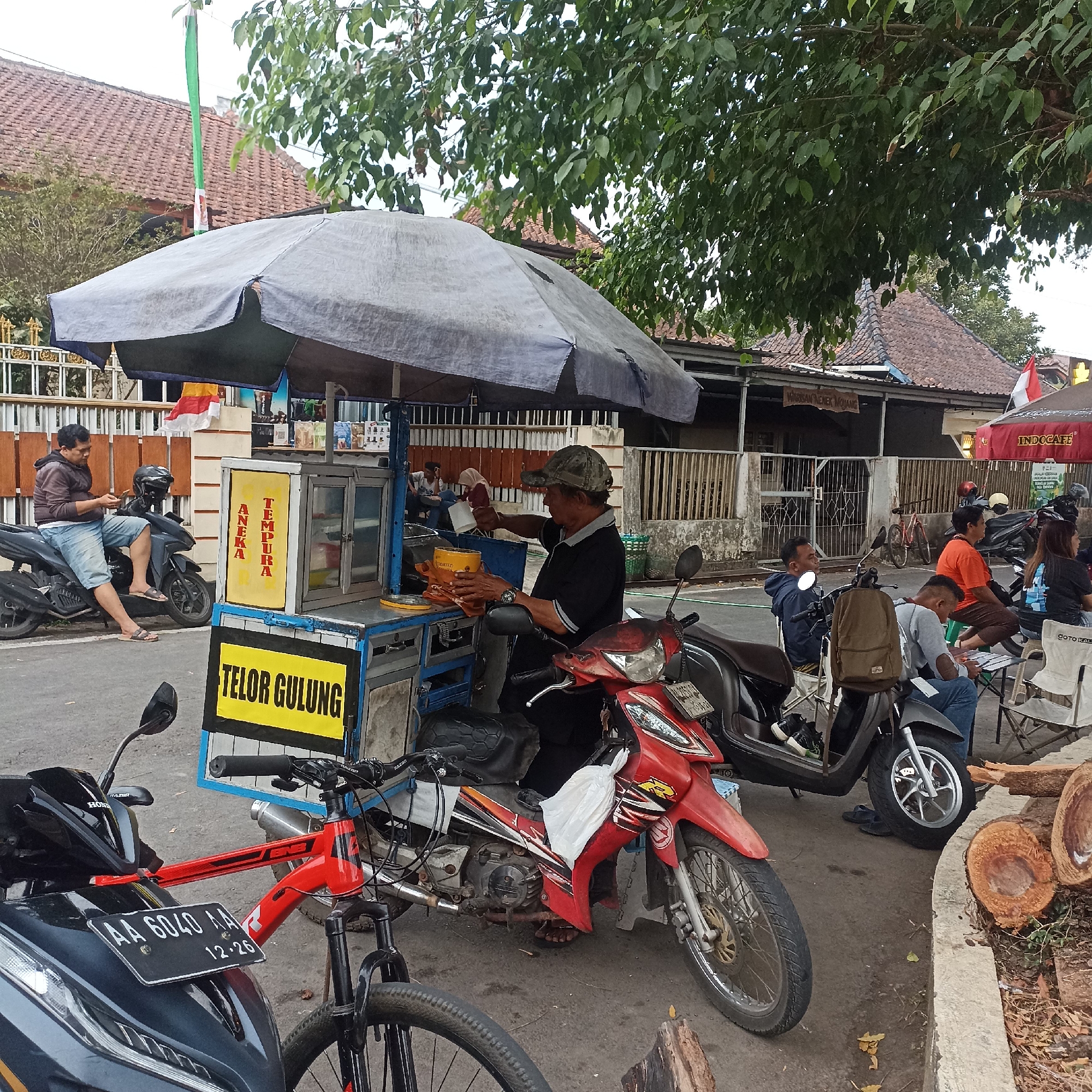 In Magelang, Indonesia, an outdoor street scene unfolds under a large umbrella providing shade to a food cart. A man in a black shirt and camouflage cap is actively preparing food at the cart, holding a yellow cup and a brown cloth. The cart itself is decorated with a yellow sign that reads "TELOR GULUNG" (rolled egg) and another vertical sign with "ANEKA" and "TEMPURA".
Several motorcycles are parked around the cart. A man in a maroon hoodie and denim shorts sits on one of the motorcycles, looking at his phone. Another motorcycle, a red one, is positioned in front of the cart, with a white plastic bag draped over its handlebars. A bicycle with a red frame is parked beside the red motorcycle.
In the background, people are seated on folding chairs near another stall, one of whom is holding a white cup. A red and white Indonesian flag is visible on a pole. The environment includes buildings with tiled roofs and a fence, suggesting a residential or commercial area. Logs are stacked on the right side of the image, near a concrete curb. The ground is paved with asphalt.