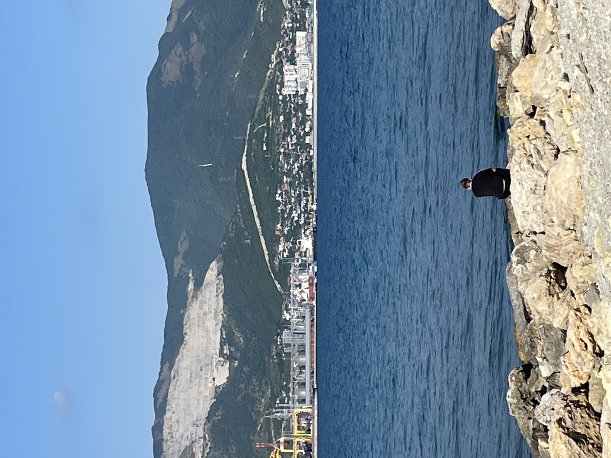 A man is sitting on rocks by the sea in Sheptal'skii, Russia, facing the water. In the distance, a city lines the coast, with buildings and industrial facilities visible. Behind the city, a large mountain range rises, its slopes covered in trees and a large, white quarry. The sky is clear and blue with a single cloud.