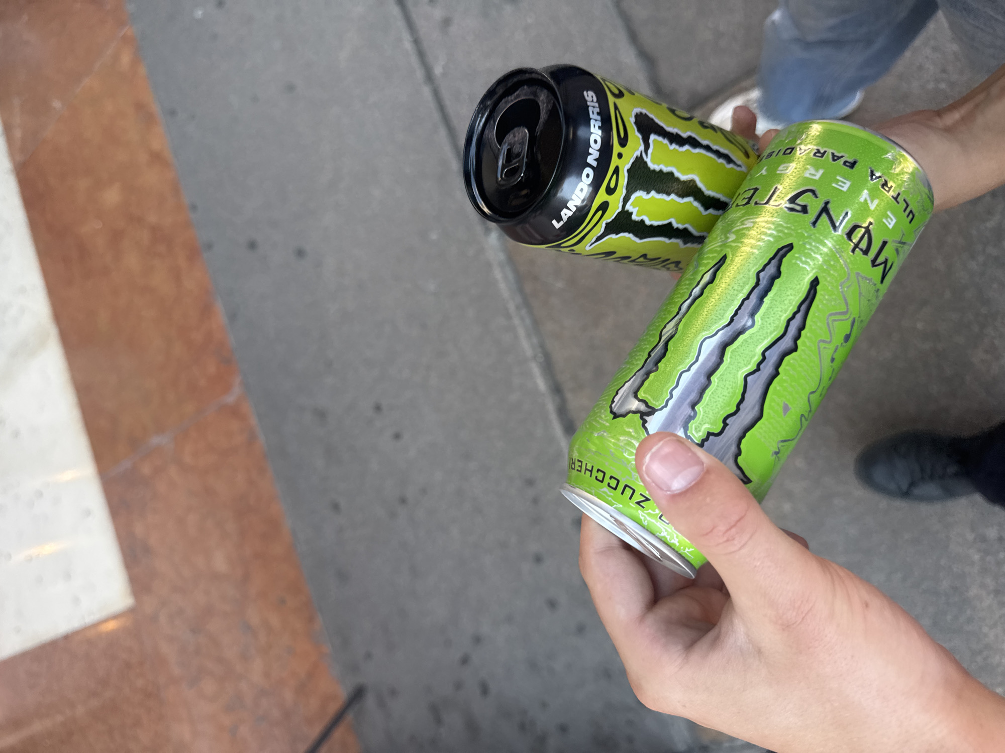 In Milan, Italy, a person's hands hold two Monster Energy cans outdoors. One can, a "Lando Norris" edition, is black and lime green. The other, a lime green "Ultra Paradise" flavor, is being held closer to the camera, with text partially visible. The background shows a grey concrete sidewalk and a reddish-brown and white tiled surface, possibly part of a building's exterior or a plaza.