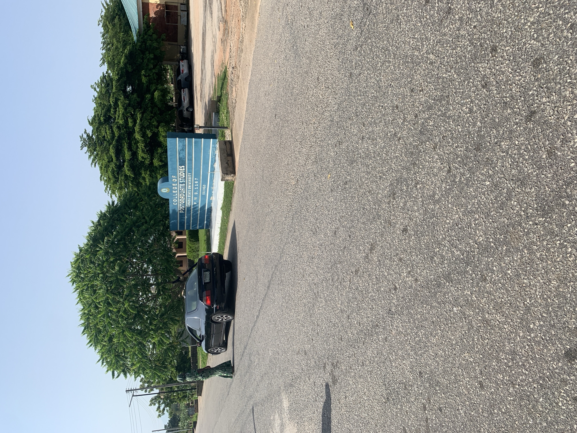 The image depicts an outdoor scene on a paved road, likely in Jalingo, Nigeria. In the foreground, the asphalt road surface is visible, with some scattering of leaves. To the left, a person wearing a green patterned traditional outfit stands facing away from the camera, partially blocking the view of a black sedan parked on the side of the road. The car is facing towards the right of the frame.
Further to the right, a blue sign stands, displaying text related to an educational institution. The sign reads "COLLEGE OF POSTGRADUATE STUDIES", "TARABA STATE UNIVERSITY", "P. M. B. 1167", and "JALINGO". Behind the sign, several parked cars are visible in a lot. A building with a green roof and red brick accents is also present in the background. Two large, lush green trees dominate the mid-ground, with one exceptionally large tree to the left and another to the right. The sky above is clear and blue, indicating a daytime scene. The overall environment appears to be a campus or institutional setting.