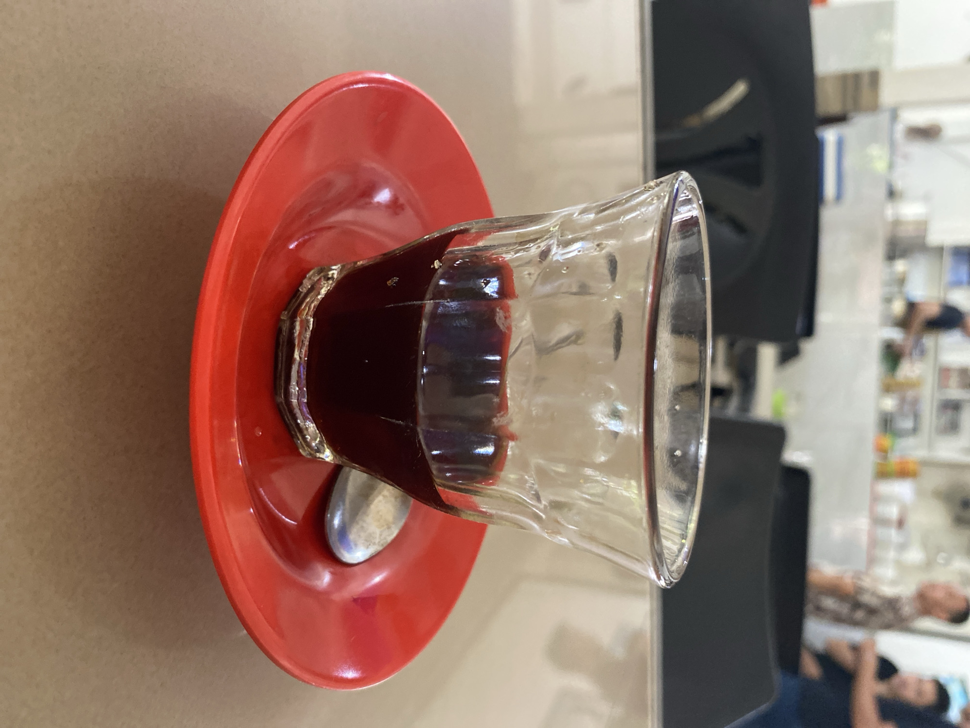 A close-up, eye-level shot features a small glass of dark liquid, likely coffee, resting on a bright red saucer. The glass is transparent with a textured surface, and the liquid fills about half of it. A small, silver spoon is positioned on the saucer to the right of the glass. The saucer is slightly curved, creating a reflective surface. The background is blurred, showing elements of an indoor setting that appears to be a cafe or restaurant. In the distance, out-of-focus figures can be seen, including one person wearing a patterned shirt and another in a dark shirt. This scene is situated in a cafe in Lhokseumawe, Indonesia.