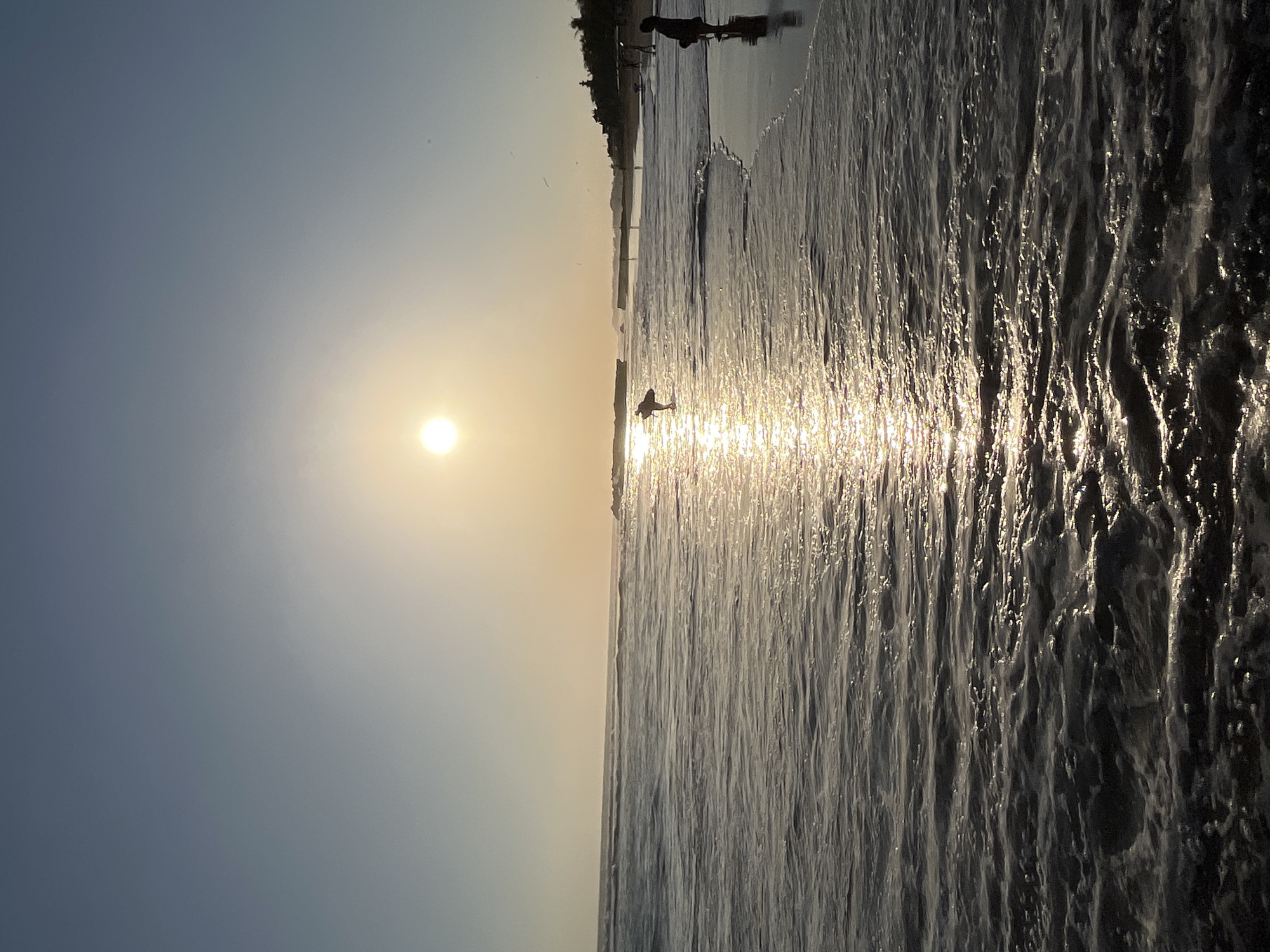 This is a medium shot, eye-level, horizontal photograph of a beach in North Kuta, Indonesia, during sunset. The sun, a bright, out-of-focus orb, is positioned in the upper center of the frame, casting a golden glow across the water. The sky transitions from a pale yellow near the horizon to a light blue at the top of the image. The ocean fills the lower two-thirds of the frame, with gentle waves rolling towards the shore. The water is reflective, with a shimmering path of sunlight stretching from the horizon towards the foreground.
In the middle ground, a lone surfer stands in the shallow water, silhouetted against the sun and holding a surfboard. Further down the beach, a person is standing on the wet sand, facing the ocean. The shoreline is lined with palm trees and structures, suggesting a developed coastal area. The overall mood is serene and tranquil, capturing the beauty of a tropical sunset.