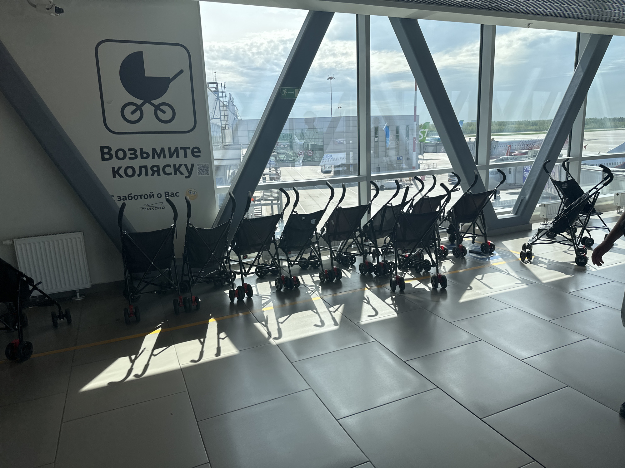 The image shows a row of black strollers lined up against a large window in Pulkovo Airport in Saint Petersburg, Russia. A sign in Russian with a stroller graphic, translated roughly to "Take a stroller, with care for you, Pulkovo," is visible. Airport infrastructure and airplanes are seen through the window. The floor is tiled, and bright sunlight casts shadows of the strollers on it.