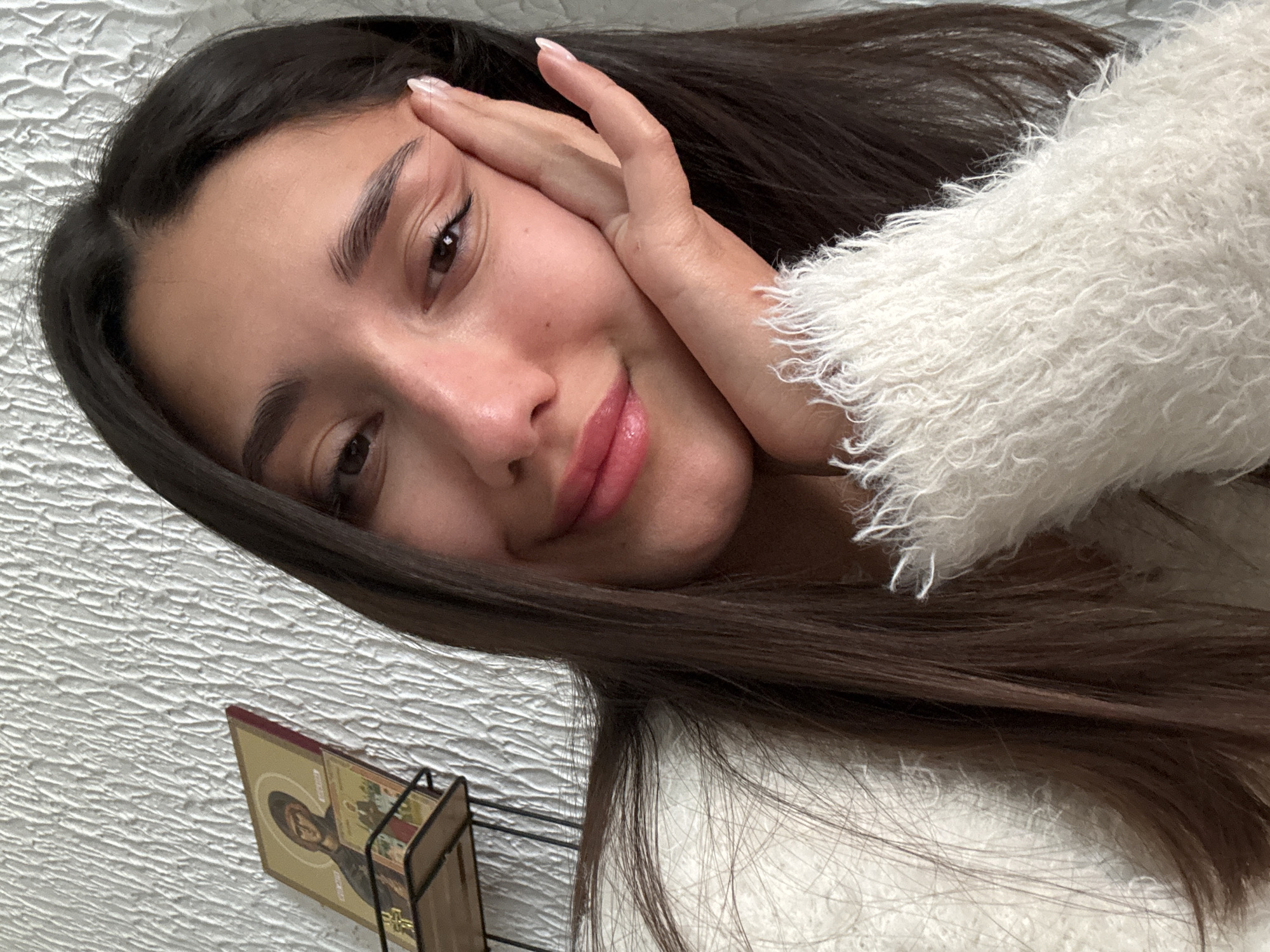 The image displays a close-up indoor portrait of a young adult female, with the user-provided location being Sutomore, Montenegro. The subject is centrally positioned, looking directly into the camera with a slight smile. Her long, dark brown hair is parted slightly off-center, cascading over her left shoulder and chest, partially obscuring a white, fuzzy, long-sleeved sweater. Her left hand, with light-colored manicured fingernails, rests against her left cheek. Her lips are adorned with a glossy pink or nude application. The background is a white or off-white wall with a distinctive textured finish. To the upper left, a black metal stand supports two religious icons; the upper icon depicts a haloed figure in an Orthodox Christian style on a gold and red panel, with a smaller icon visible beneath it. The scene is illuminated by soft, even lighting.