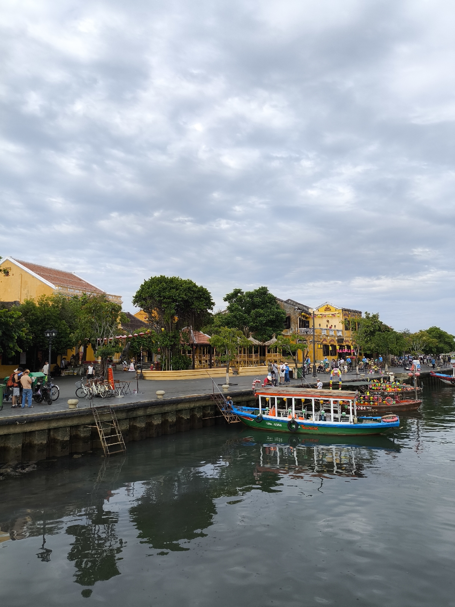 The image depicts a waterfront scene in Hoi An, Vietnam, likely along the Thu Bon River. The foreground shows the dark, rippling water with reflections of the sky and surrounding structures. A green and blue boat with an orange roof, identified by the hull number "QNA.0358," is moored near the shore. Behind the boat, a brown boat with numerous colorful decorations is also docked.
The middle ground features a stone embankment and a paved walkway lined with a railing. Several people are visible on the walkway, some standing and conversing, others walking or cycling. Various bicycles and motorbikes are parked along the walkway. In the background, traditional Vietnamese architecture dominates, characterized by yellow-painted buildings with terracotta roofs, interspersed with lush green trees and some thatched-roof structures. The sky is overcast with scattered clouds.