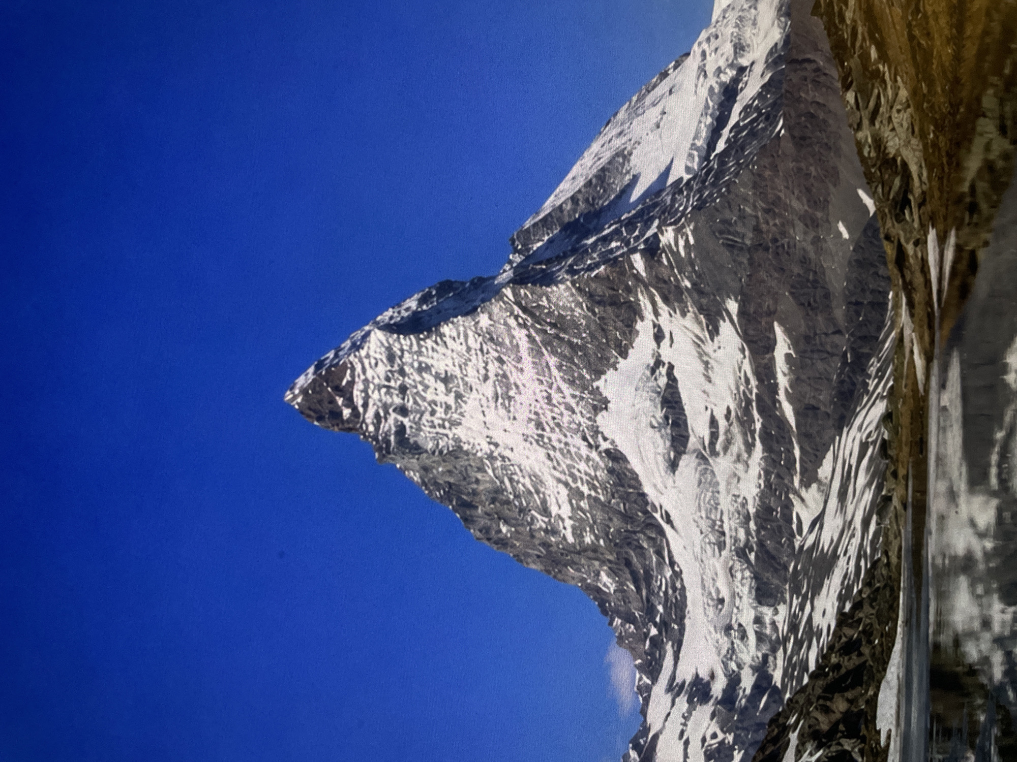 A towering, snow-capped mountain dominates the center of the frame, its peak sharply defined against a clear, vibrant blue sky. The mountain's slopes are a rugged mix of dark rock and bright white snow, with visible glaciers and scree. In the foreground, a calm body of water reflects the majestic mountain, creating a mirrored image. The water's surface is disturbed by ripples in some areas. The surrounding terrain is rocky and barren, with patches of dry grass and small snowdrifts. The overall scene depicts a dramatic and awe-inspiring natural landscape.