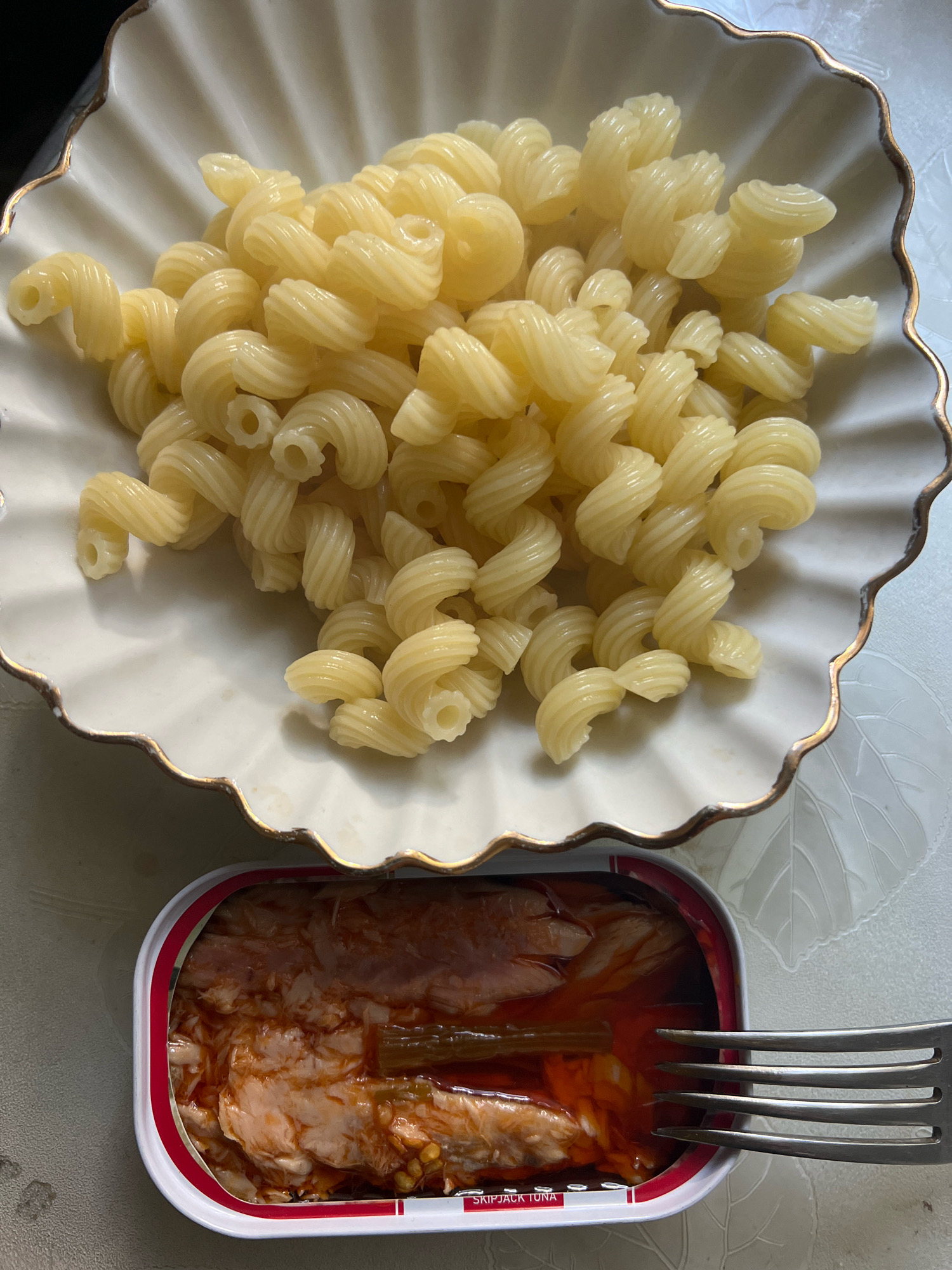 A close-up, overhead shot shows a white, scalloped bowl filled with cooked fusilli pasta. To the right of the bowl, a fork is placed in an open can of tuna. The tuna is flaky and mixed with a red sauce. The background is a light-colored surface with a subtle leaf pattern. There are no people visible in the image.