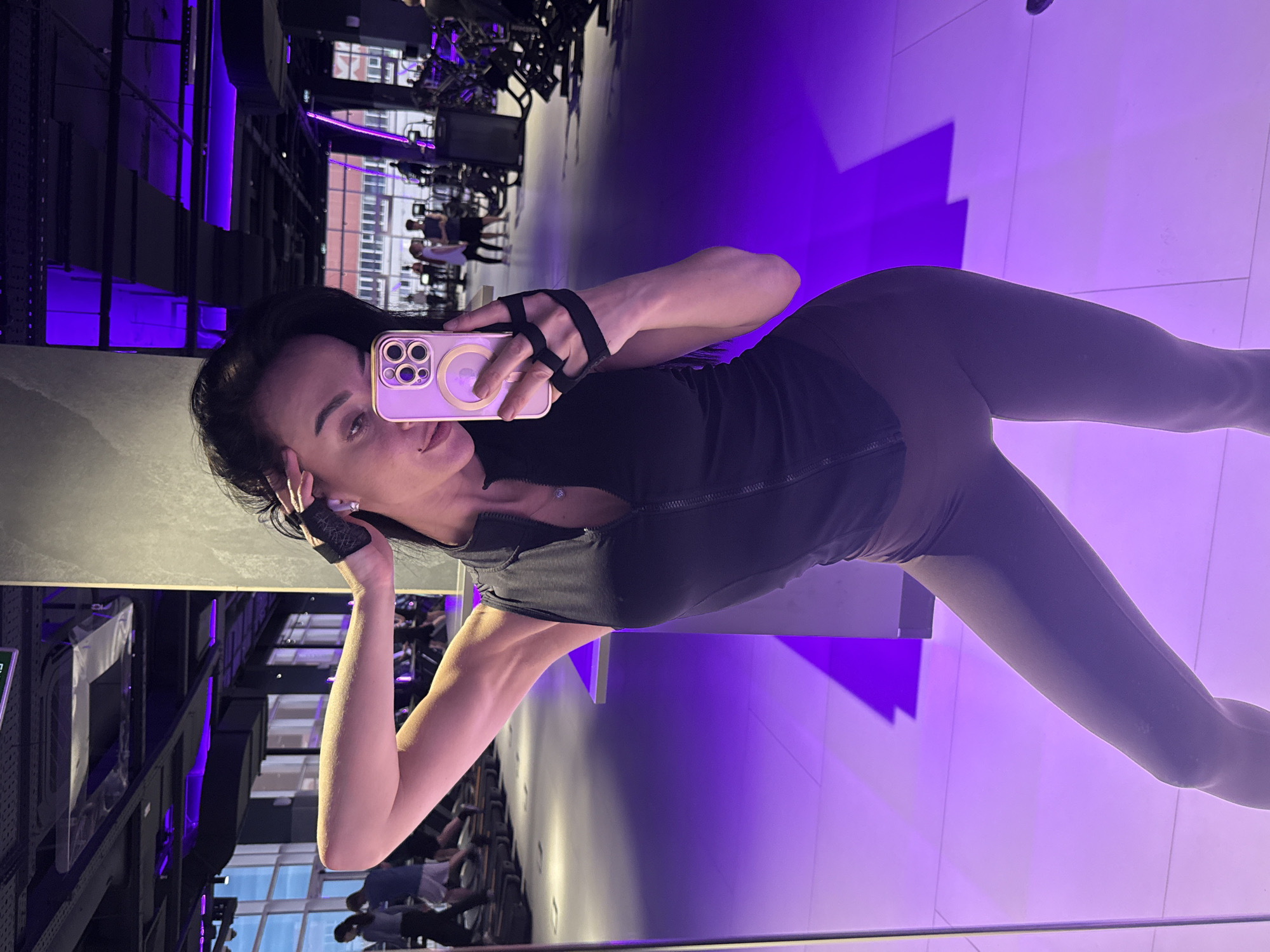 A woman is standing in what appears to be a gym, taking a selfie using a phone with a pale pink case. She is wearing a dark-colored workout outfit consisting of a sleeveless top and leggings, and a black wrist support is on her hand that is holding the phone. Her other hand is near her ear. The gym environment includes exercise equipment, a mirrored wall reflecting the scene, and large windows revealing the exterior. The lighting in the gym is a mix of natural and artificial with distinct purple highlights. The location is Krasnoyarsk, Russia.