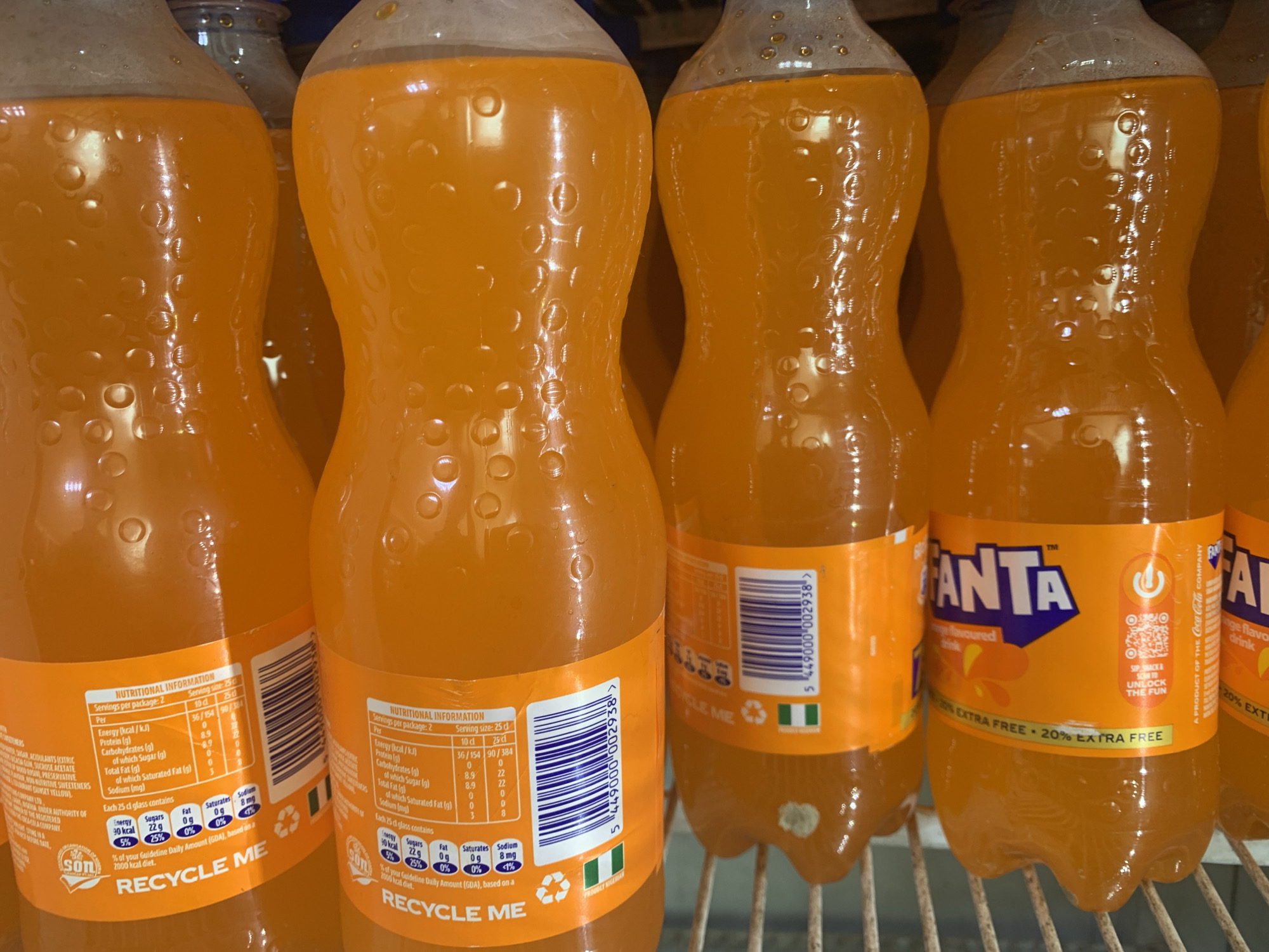A row of bright orange soda bottles is displayed on a metal shelf. The bottles are clear plastic and have textured surfaces with small indentations. Each bottle has a vibrant orange label with white and blue text. The labels show nutritional information, barcodes, and the Fanta logo. Some bottles have plastic caps that are light gray. The lighting is bright, reflecting off the shiny surfaces of the bottles and the liquid inside. The background is dark, suggesting a refrigerator or cooler.