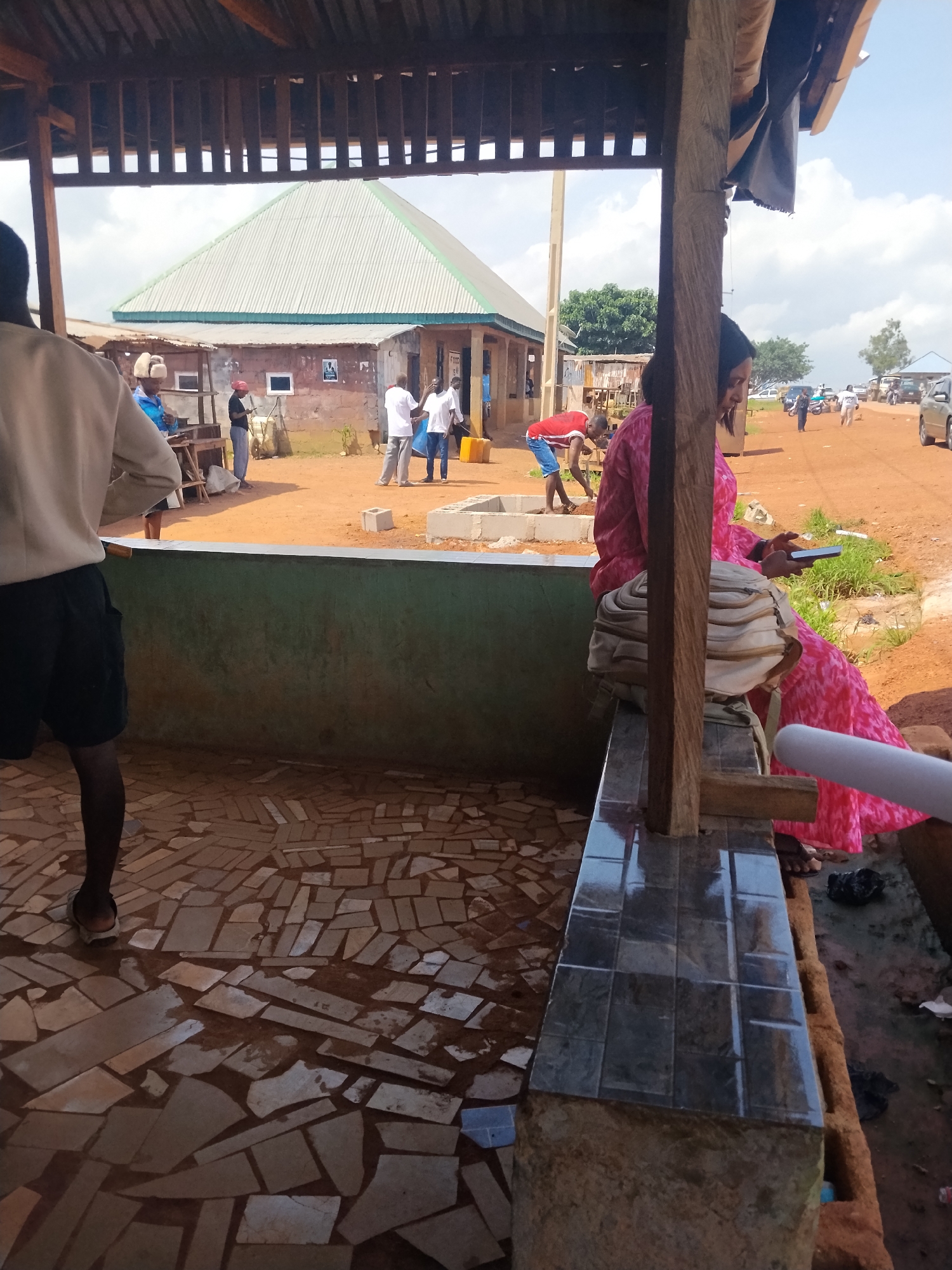 In Durumi, Nigeria, an outdoor scene unfolds under a shaded structure with a corrugated metal roof supported by wooden beams. The foreground is partially occupied by a person from behind, wearing a light-colored long-sleeved shirt and black shorts. They are standing on a floor paved with irregular, light-colored tiles.
To the left, a building with a green trim and a light-colored, ridged roof is visible. In front of it, several people are engaged in various activities. Three men in white shirts and dark pants stand together, and another man in a blue shirt is near a wooden structure. One individual, wearing a red shirt and blue shorts, is bending over, possibly working on the ground.
Further in the background, a dirt road stretches out, with vehicles, including cars and motorcycles, parked or moving. A woman in a vibrant pink patterned dress is seated to the right, looking down at a dark-colored mobile phone she holds in her hand. A beige backpack rests beside her. The ground is predominantly reddish-brown earth, with some patches of sparse vegetation. The sky is cloudy.