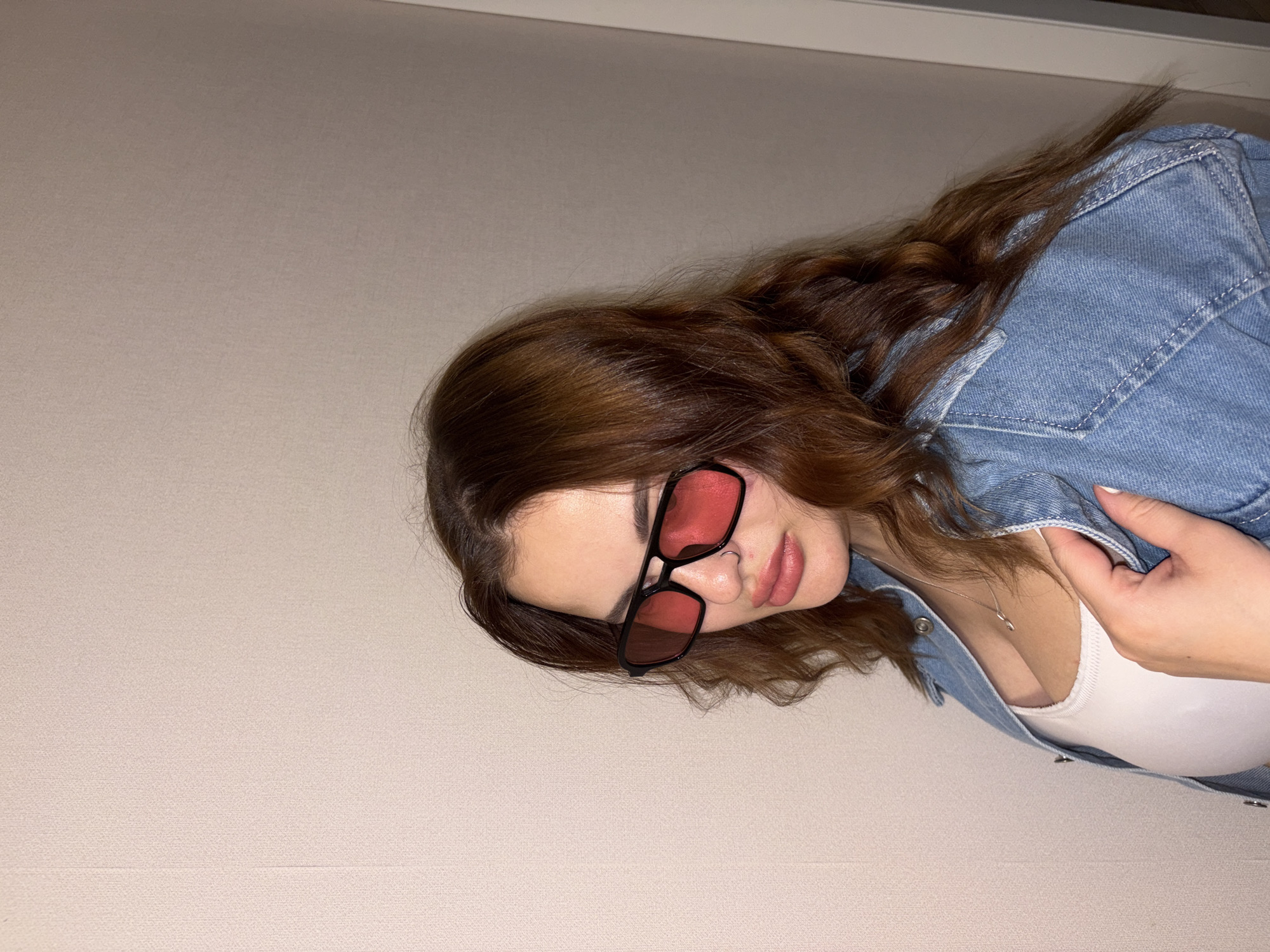 A close-up portrait shows a young woman posing indoors against a plain, light beige wall. She has long, wavy, reddish-brown hair cascading around her shoulders. Her head is tilted slightly, and she is wearing black-framed sunglasses with distinct red-tinted lenses that obscure her eyes. Her lips are a natural pinkish hue, slightly parted, and she has a small hoop piercing on her right nostril. She is dressed in a light blue denim jacket over a white top, with a delicate silver necklace visible around her neck. One of her hands, with white-painted fingernails, is casually holding the lapel of her denim jacket. The lighting is soft and even, suggesting an indoor setting. There is no visible text in the image. The scene is set in Krasnodar, Russia.