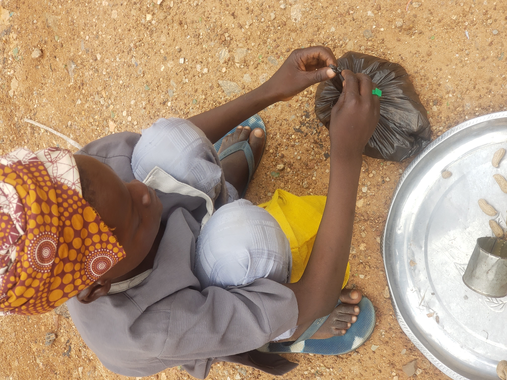 A person kneels on sandy ground in Narayi, Nigeria, wearing a gray tunic and light blue checkered pants. They have a patterned head covering. In front of them is a large silver platter containing peanuts and a small metal cylinder. The person is holding a black plastic bag and tying it shut with their hands. To their left, a yellow fabric is visible. The ground is composed of sand and small stones.