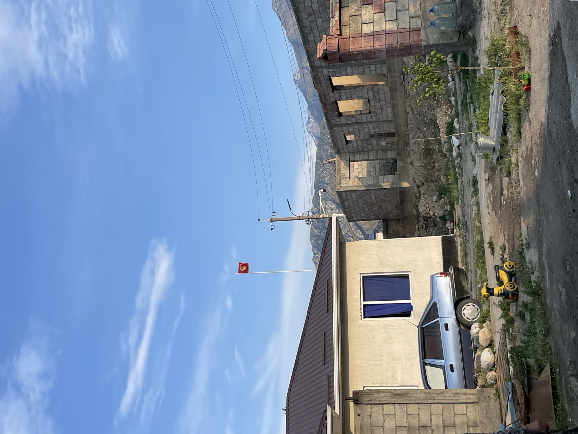 The image depicts a street scene in Ak-Sai, Kyrgyzstan. A light blue sedan is parked in front of a beige building with a dark brown metal roof. To the right of the building, the skeletal remains of a brick structure under construction are visible. In the background, majestic mountains form a backdrop under a clear blue sky with scattered clouds. A flag with a yellow sun and a tündük (a yurt crown) on a red field, representing Kyrgyzstan, flies from a tall flagpole. A yellow toy ride-on car is positioned on the ground in front of the parked car. The overall impression is one of a developing residential area with a clear view of the natural landscape.