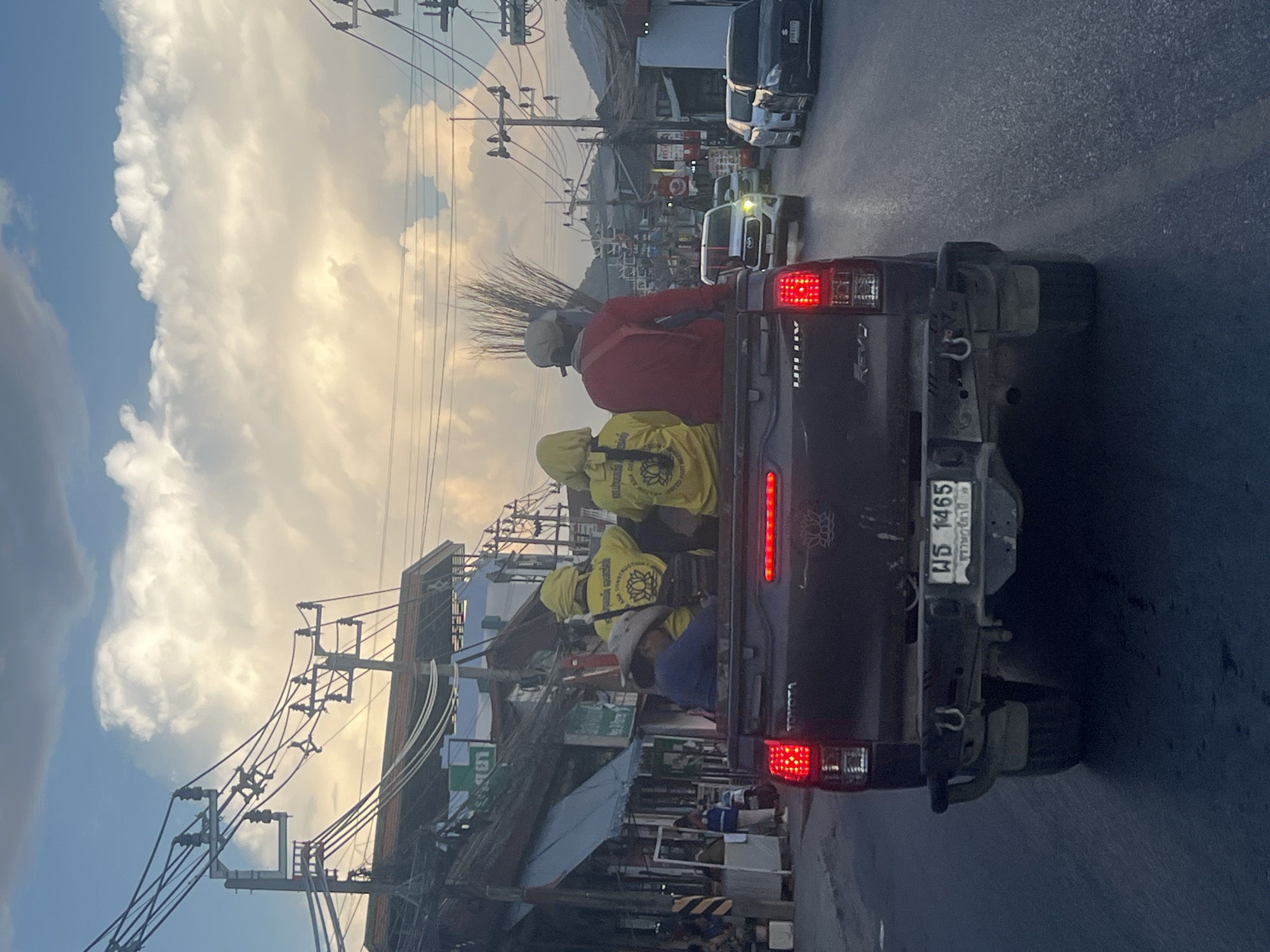 In Ko Samui District, Thailand, a dark-colored pickup truck with its rear facing the camera drives on a paved road. The truck bed is occupied by four individuals, all facing away from the camera. Three of them are wearing yellow shirts and hoods; one has a lotus logo with text that appears to be "ASIA CONSTRUCT" and "PROMOTED PART". The fourth individual in the truck bed wears a red shirt and a beige cap, and is holding a broom. In the passenger seat of the truck bed, a person wearing a blue shirt and a wide-brimmed hat is visible. The truck has a license plate with Thai characters and the number "WS 1465". The surrounding environment shows buildings with awnings, utility poles with numerous wires, and other vehicles on the road. The sky above is filled with dramatic clouds and a soft, warm light suggesting either sunrise or sunset.
