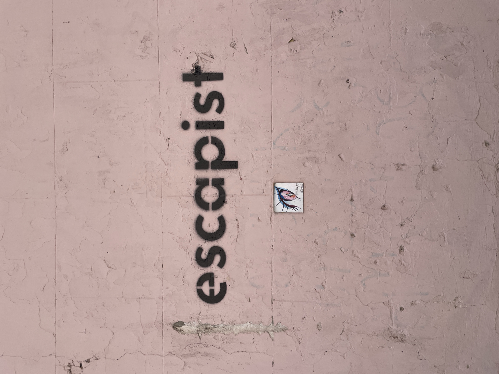 A pink exterior wall features vertical seams, visible layers of paint and a horizontal fissure. The word "escapist" is stenciled in black vertically on the left. To the right of the text is a small square art tile. The tile depicts an eye with red and blue colors. The location is Tbilisi, Georgia.