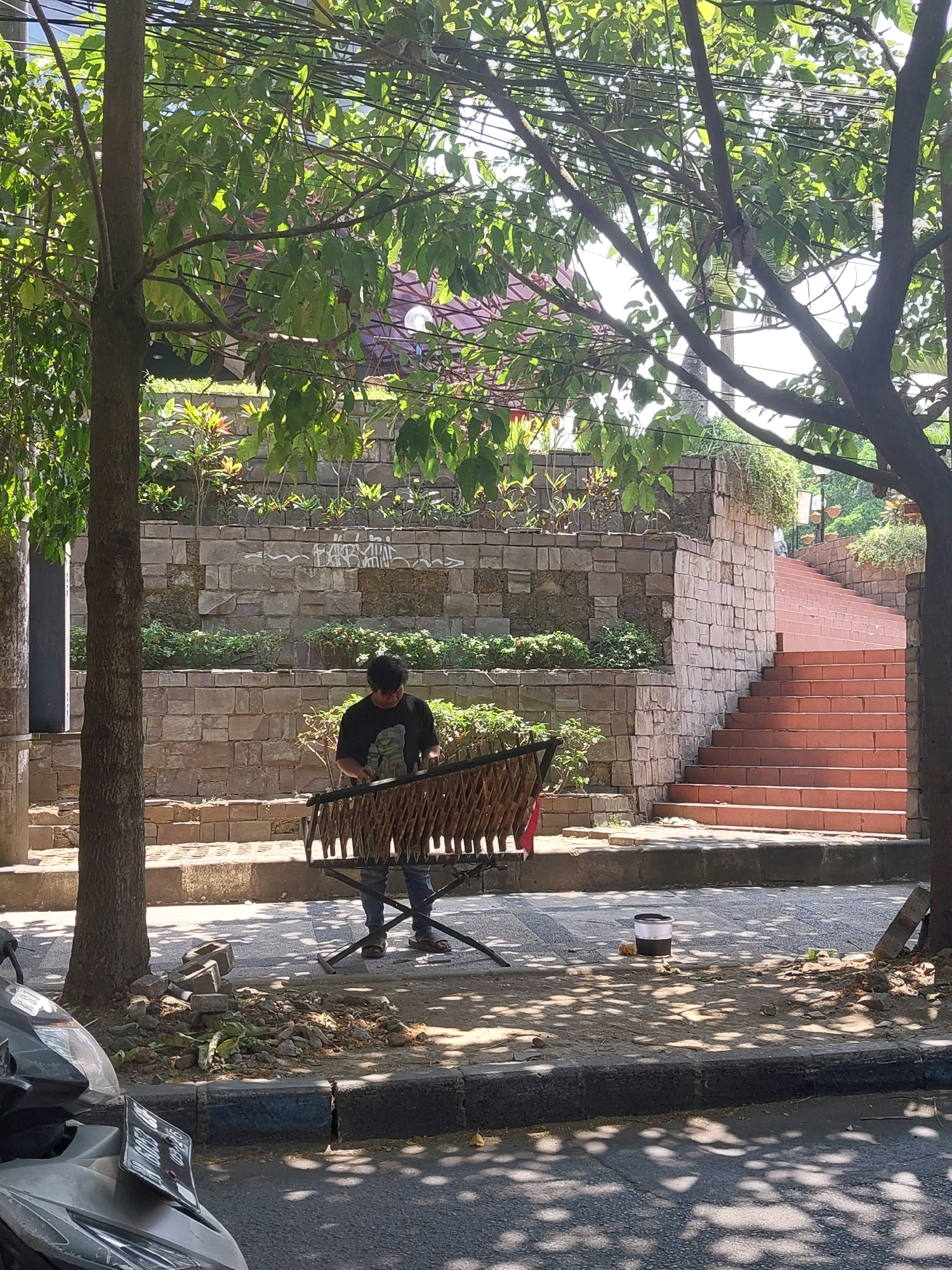 A man is standing next to a musical instrument on a sidewalk in Malang, Indonesia. The man is wearing a black t-shirt and blue jeans. The instrument appears to be a type of xylophone or gamelan instrument, with many wooden bars arranged in rows. The sidewalk is paved with cobblestones, and there is a tree with green leaves overhead, casting shadows on the ground. Behind the man and the instrument is a terraced stone wall with plants growing on it. To the right of the stone wall are a set of red brick stairs leading upwards. In the foreground, a motorcycle is parked on the street. A cylindrical object with a white top and black base is on the sidewalk near the stairs. The overall scene is lit by bright sunlight.