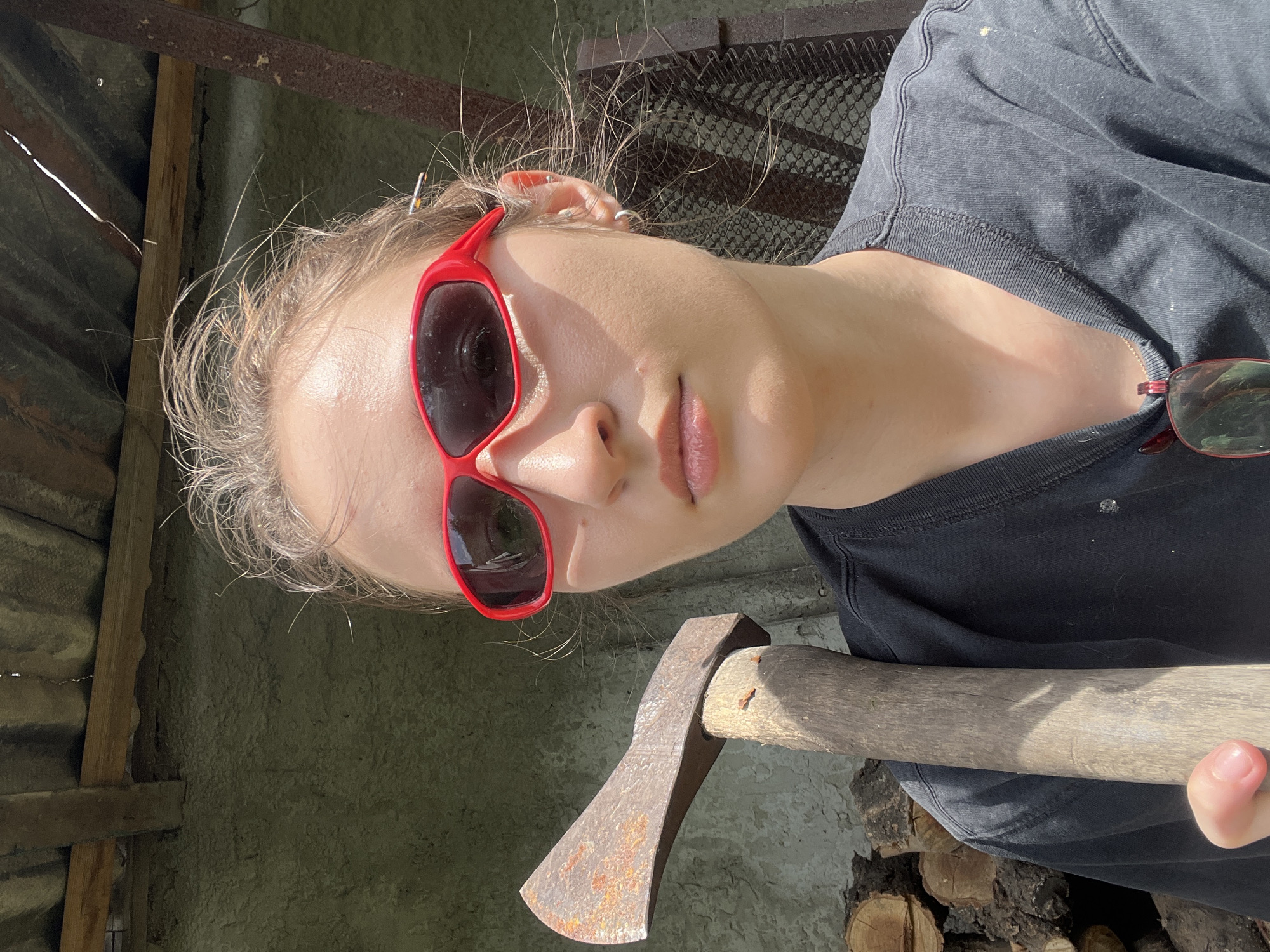 An outdoor, waist-up, self-portrait shows a young woman holding a rusted axe with a wooden handle. She is wearing red-framed sunglasses, a dark t-shirt, and a silver hoop earring in her left ear. Her hair is light brown and appears windswept. Behind her, a stack of cut logs is visible, along with a gray, textured wall and a corrugated metal roof supported by wooden beams. The scene appears to be in a rural or backyard setting.