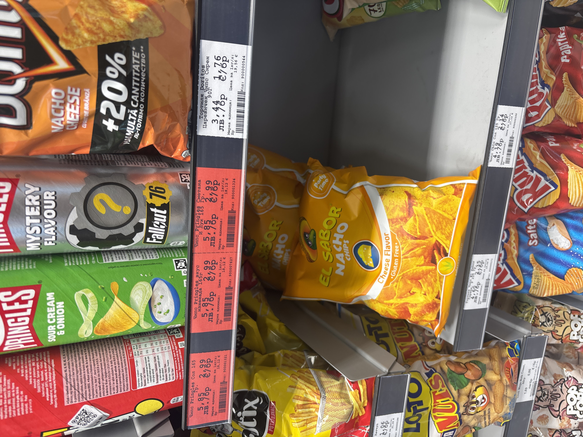 The image depicts a shelf of potato chips in a store, likely in Plovdiv, Bulgaria, judging by the Cyrillic text on the price labels. The chips are stacked vertically, with various brands and flavors visible. From left to right, the brands include Pringles (Sour Cream & Onion, Mystery Flavor, and Creamy), Doritos (Nacho Cheese), El Sabor (Nacho Chips - Cheese Flavor), Chio (Salted), and another variety with paprika flavor. The price labels indicate prices in Bulgarian Lev (лв.) and Euro (€). The shelf is gray, and there is a slight downward angle to the view. There are other snack items further down on the shelf, including peanuts. No people are visible in the frame.