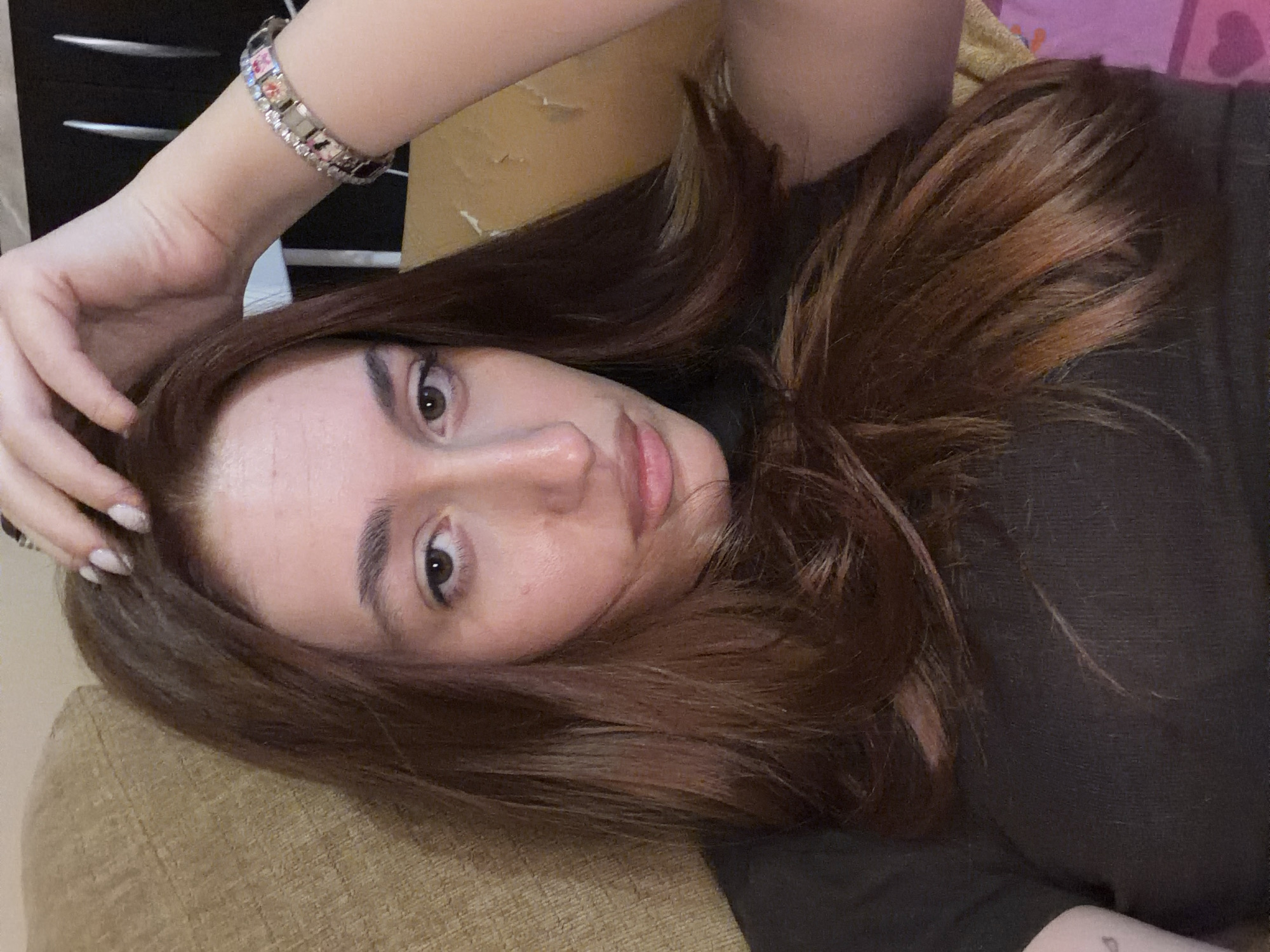 A woman with reddish-brown hair is lying down, possibly on a couch or bed. She is wearing a dark-colored shirt and a bracelet. Her left hand is near her head. The background includes a light brown surface and a dark cabinet with drawers. There's no indication of location in Plovdiv, Bulgaria.