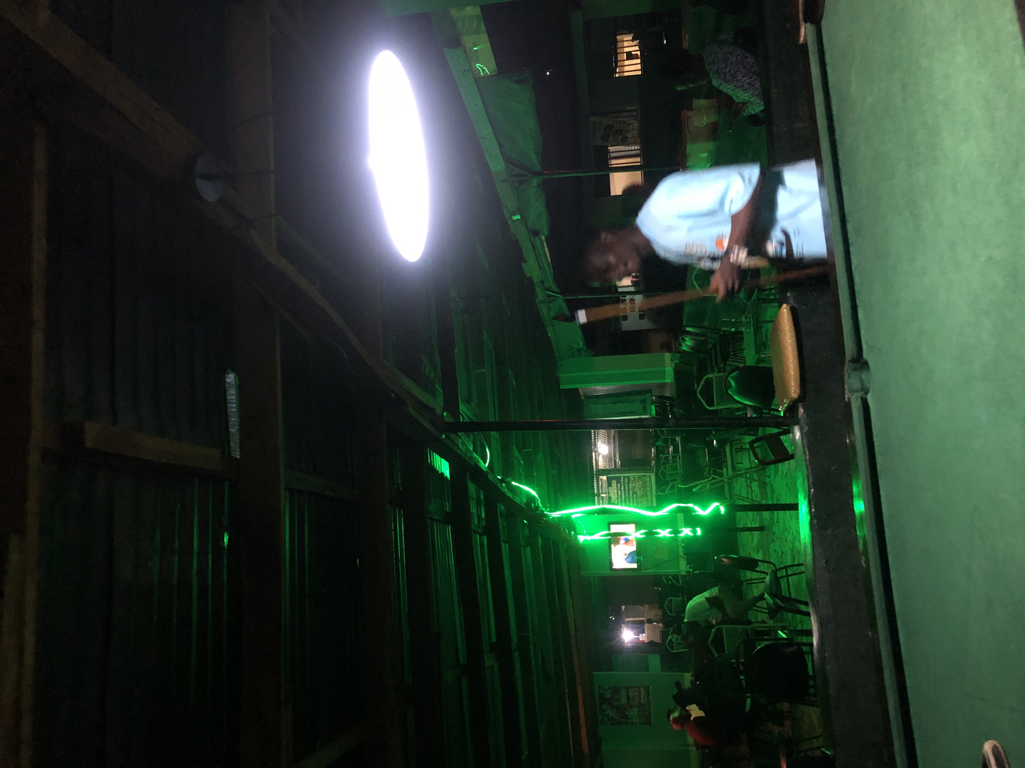 A dimly lit indoor establishment, possibly a bar or a casual gathering spot, is captured from a low angle, focusing on a pool table in the foreground. The scene is dominated by an intense green ambient light, emanating from string lights that snake across the ceiling and walls, and also from what appears to be a laser light effect. A bright, large, oval-shaped light fixture hangs from the ceiling in the mid-ground, casting a strong white light that contrasts with the green.
In the mid-ground, a young Black man wearing a light blue t-shirt is the primary subject, holding a pool cue. He is looking to his right, slightly out of focus. To his left, in the background, other patrons are seated at tables, also dimly illuminated and partially obscured by shadows and the green light. Some chairs are visible, some stacked. A television screen on the far left displays a colorful image. In the far background, through an open doorway, a brighter light suggests another area of the establishment. The ceiling appears to be made of corrugated metal, supported by wooden beams, and there are tarps or fabric coverings suspended in some areas. The floor is not clearly visible. The overall atmosphere is casual and slightly mysterious due to the intense green lighting.