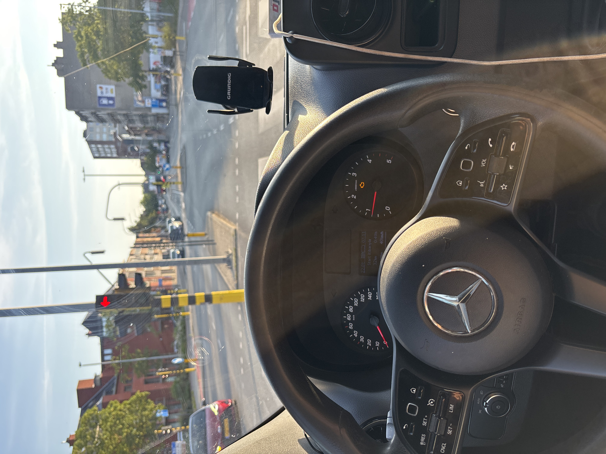 The image is taken from the driver's perspective inside a Mercedes-Benz van, looking forward through the windshield. The van is stopped at a traffic light in the city of Boom, Belgium, at the intersection of the N16 and Boom's main street, located near the local church. The van's dashboard, including the steering wheel with the Mercedes-Benz logo and airbag indicator, and the instrument cluster displaying speed and engine RPM, are visible. A Grundig phone holder is attached to the windshield. Outside, typical urban scenery of Boom is depicted, with buildings lining the street, trees, streetlights, and other vehicles. The sun is low in the sky, casting long shadows and creating a warm, golden light, suggesting it is either early morning or late afternoon. The traffic light is red, indicating the van is temporarily halted.