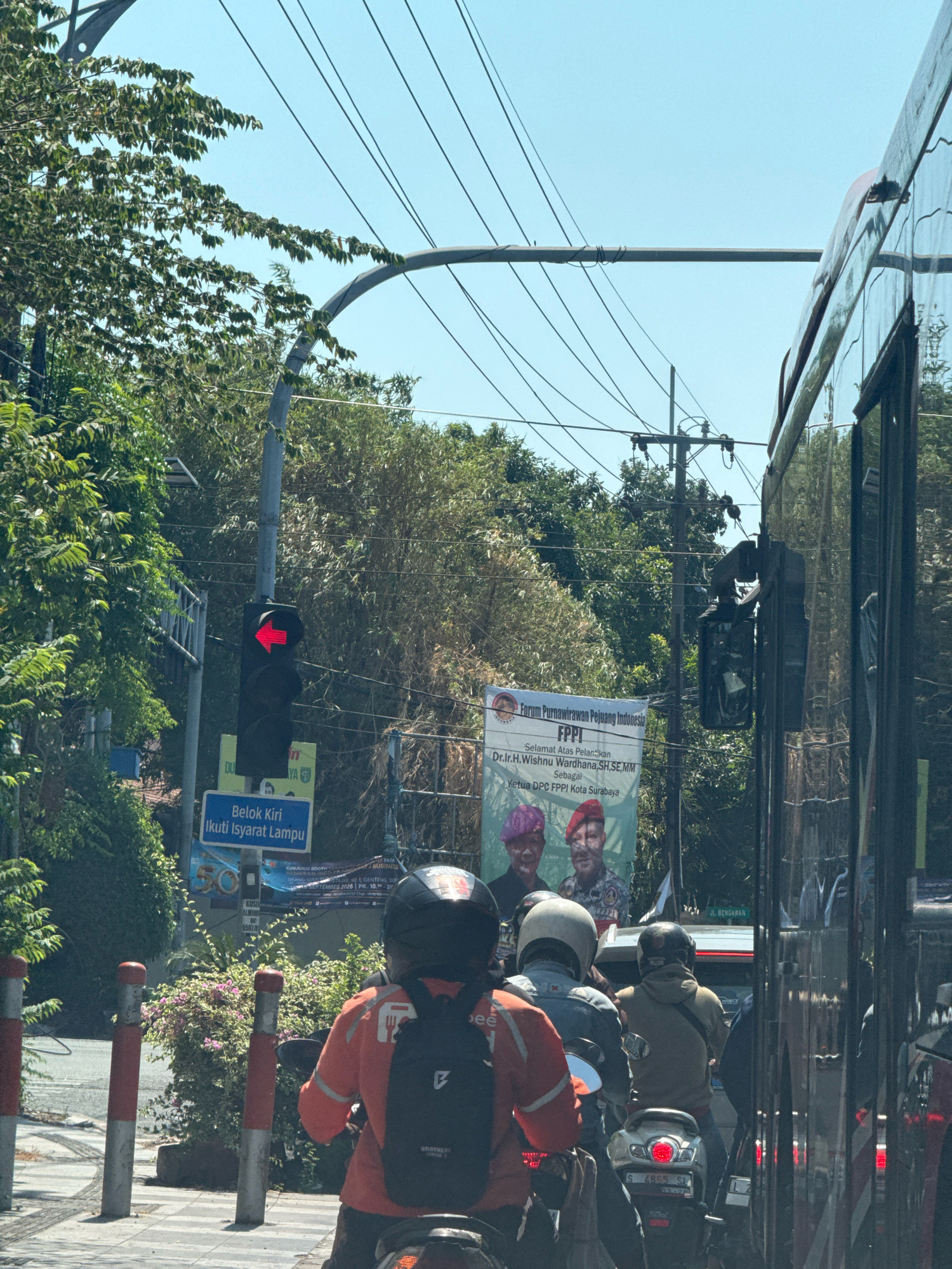 The image captures a street scene in Surabaya, Indonesia, with a focus on traffic and a traffic light. A red arrow on the traffic light indicates a left turn is prohibited. A sign below the traffic light reads "Belok Kiri Ikuti Isyarat Lampu", which translates to "Turn Left Follow Traffic Light Signals". Several motorcyclists are visible, some in the foreground and others further back in traffic. One motorcyclist in the foreground is wearing an orange jacket and a black backpack. In the background, a large banner is displayed with text that includes "Forum Purnawirawan Pejuang Indonesia FPPI" and "Ketua DPC FPPI Kota Surabaya", identifying an organization and its local chapter leader. Lush green trees and vegetation line the street, and a tall utility pole with wires stretches across the sky. A large vehicle, likely a bus, is reflected in the foreground, partially obscuring the view of the road behind it. The sky is clear and bright, suggesting a sunny day.