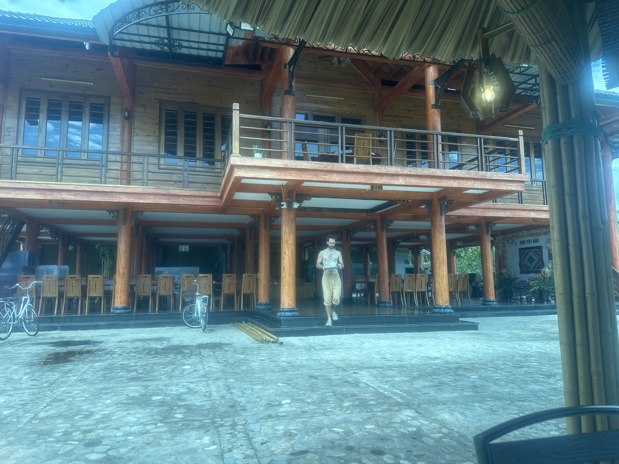 The image displays a two-story wooden building, potentially a guesthouse or restaurant, situated in Nghia Lo Ward, Vietnam. The structure features light brown wooden plank walls, contrasting with darker reddish-brown wooden pillars and beams. The second floor includes a balcony with a dark railing, fronted by multiple windows that reflect an overcast sky. A dark, ornate metal lamp with a glowing bulb hangs from a thatched roof overhang on the upper right side of the building's facade. Another smaller, wall-mounted lamp is visible on the second floor.
The ground floor presents an open-air space supported by robust wooden pillars, accessed via a black-tiled raised platform with steps. This area is furnished with numerous light-colored wooden chairs arranged around tables, indicating a dining or communal space. A sign on the back wall of the ground floor, on the right side, reads "GÓC TÂY BẮC".
An adult male, shirtless and wearing light tan shorts, with short dark curly hair and a beard, is depicted in motion, descending the steps from the building's ground floor. He holds a light-colored bowl or tray containing a cup in both hands.
In the paved foreground, two bicycles are parked on the left. A stack of several bamboo poles lies horizontally on the ground near the base of the steps. The ground surface is a textured concrete or stone pavement with several visible wet patches. A large bamboo pillar is partially visible on the far right, and a dark chair back is present in the extreme lower right foreground. The overall scene suggests a daytime setting with diffused natural light.