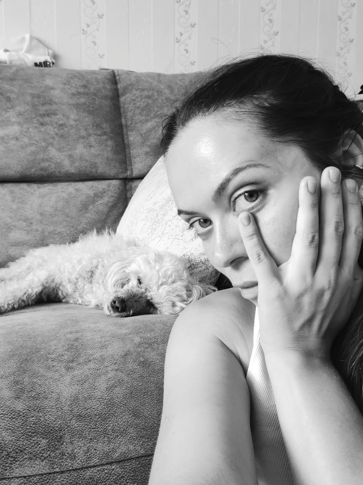 In a close-up, black and white selfie, a woman with dark hair pulled back rests her right hand on her face, obscuring part of her cheek and eye. Her gaze is directed slightly away from the camera, towards the left side of the frame. To her left, a small, white, curly-haired dog sleeps on a grey sofa. The sofa is positioned in front of a wall with a floral wallpaper pattern. The background suggests an indoor setting, likely a living room, in the city of Chaikovsky, Russia.