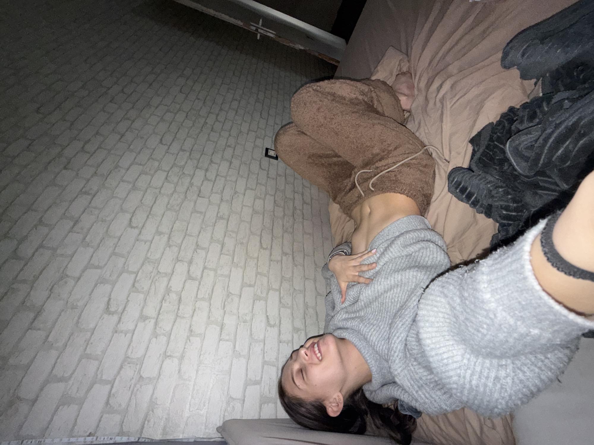 A woman is lying on a bed. She is wearing brown sweatpants and a gray sweater. Her head is resting on a pillow. She is smiling. The background includes a gray brick-like wall. The location is not identifiable as Sofia, Bulgaria.