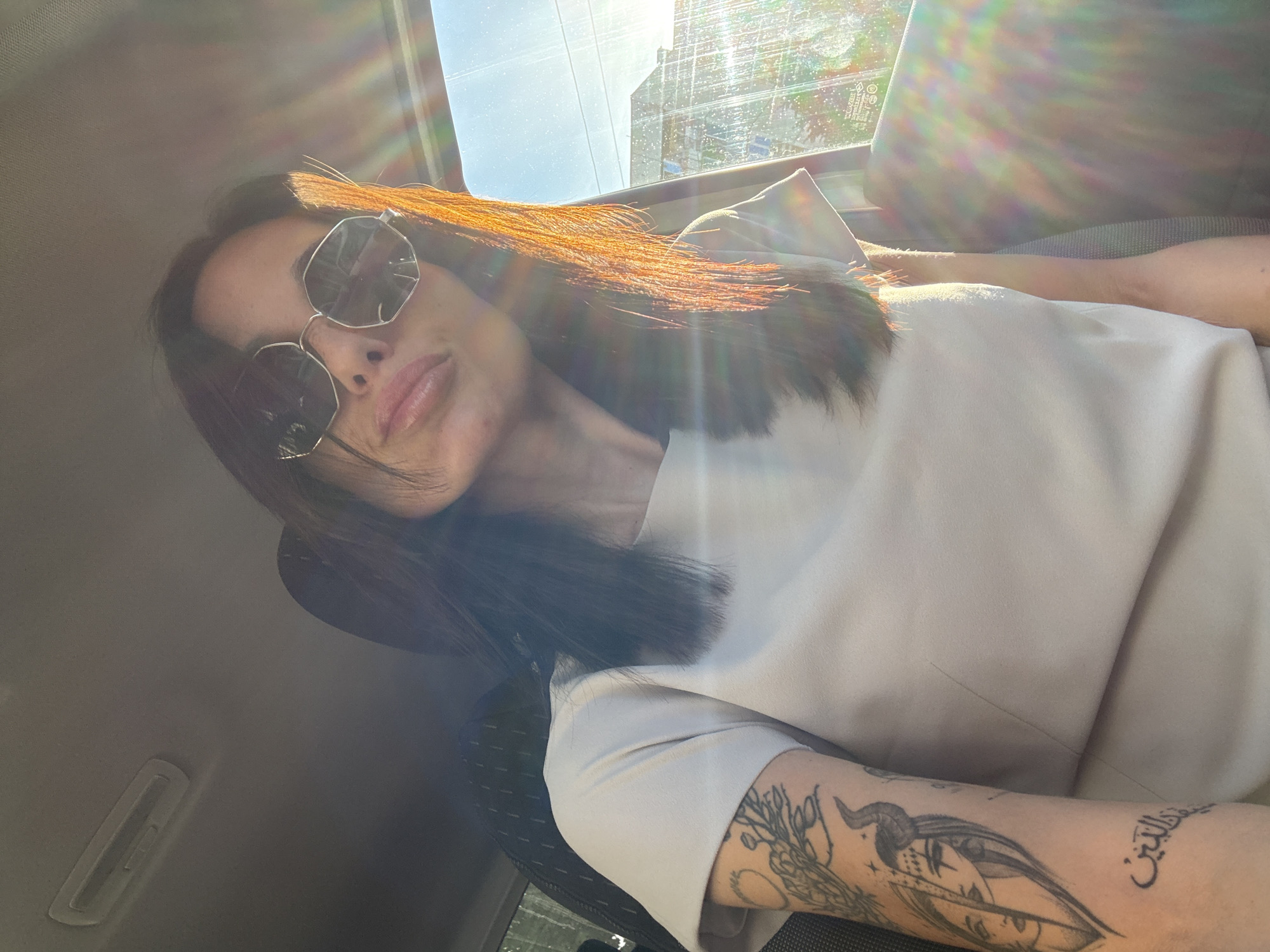 A woman with sunglasses is inside a vehicle in Pleven, Bulgaria. She has brown hair and is wearing a light-colored shirt. Her left arm has several tattoos. Sunlight is streaming in through the window. Outside, a building can be seen.