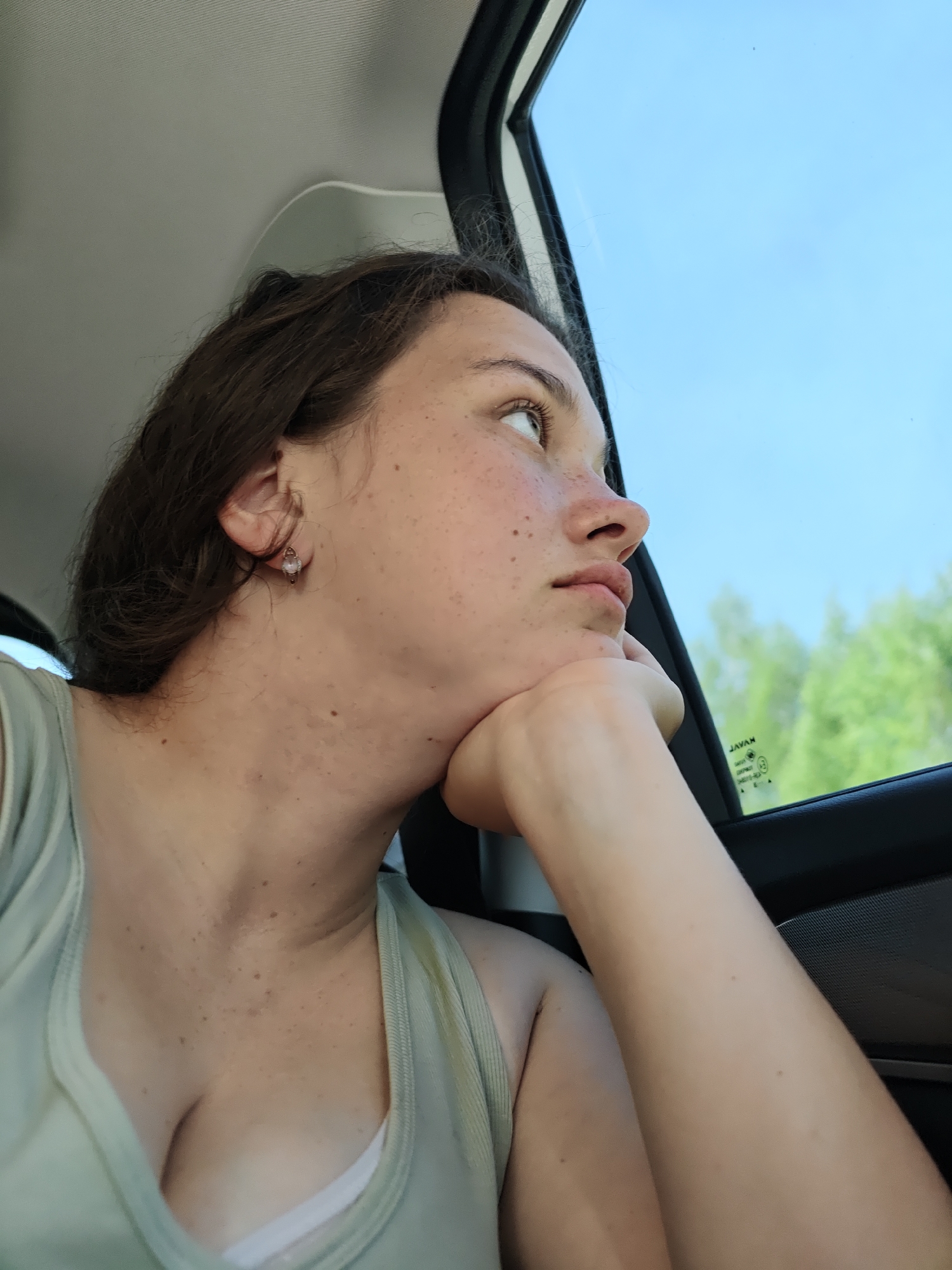 A young woman is in a car, looking out the window to the right. The window shows a clear blue sky and green trees, possibly indicating a drive through a rural area or the outskirts of a city. She is resting her head on her hand. She has brown hair and is wearing a light-colored tank top. She has freckles. The car interior is visible around her, including the window frame and part of the dashboard. There is no specific information available within the image itself to determine her precise location in Chaikovsky, Russia.