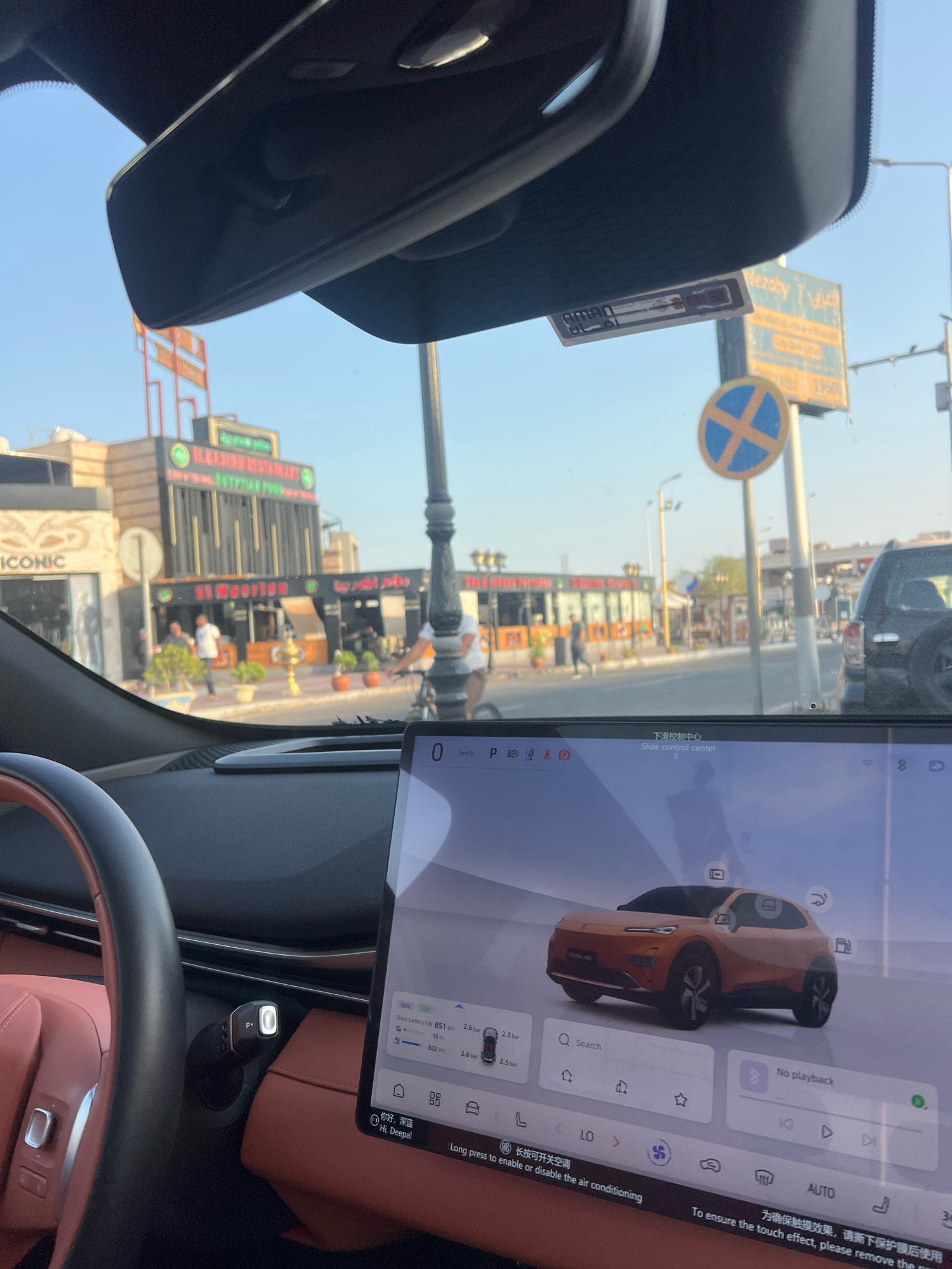 This image captures a view from inside a vehicle, looking forward through the windshield. The foreground shows the dashboard of a car with a large touchscreen display. The touchscreen displays a car icon, a search bar, and other controls. The car's interior features red leather seats.
Outside the car, a street scene unfolds. Several buildings line the street, some of which appear to be restaurants. One building prominently displays a sign that reads "EL KASHEER RESTAURANT EGYPTIAN FOOD." Another sign says "EL MOORIAN." People are visible walking on the sidewalk and in the street. A black SUV is parked on the right side of the street. Traffic signs, including a "no parking" sign, are also present. The sky is clear and blue, indicating a sunny day. The architecture and signage suggest a location in a tourist area, and the Arabic text on some of the signs confirms its Middle Eastern context. The overall impression is a snapshot of everyday life in a bustling city. Based on the identified restaurant names and architectural style, the location is likely Sharm El Sheikh, Egypt, a popular tourist destination known for its Egyptian and international cuisine.