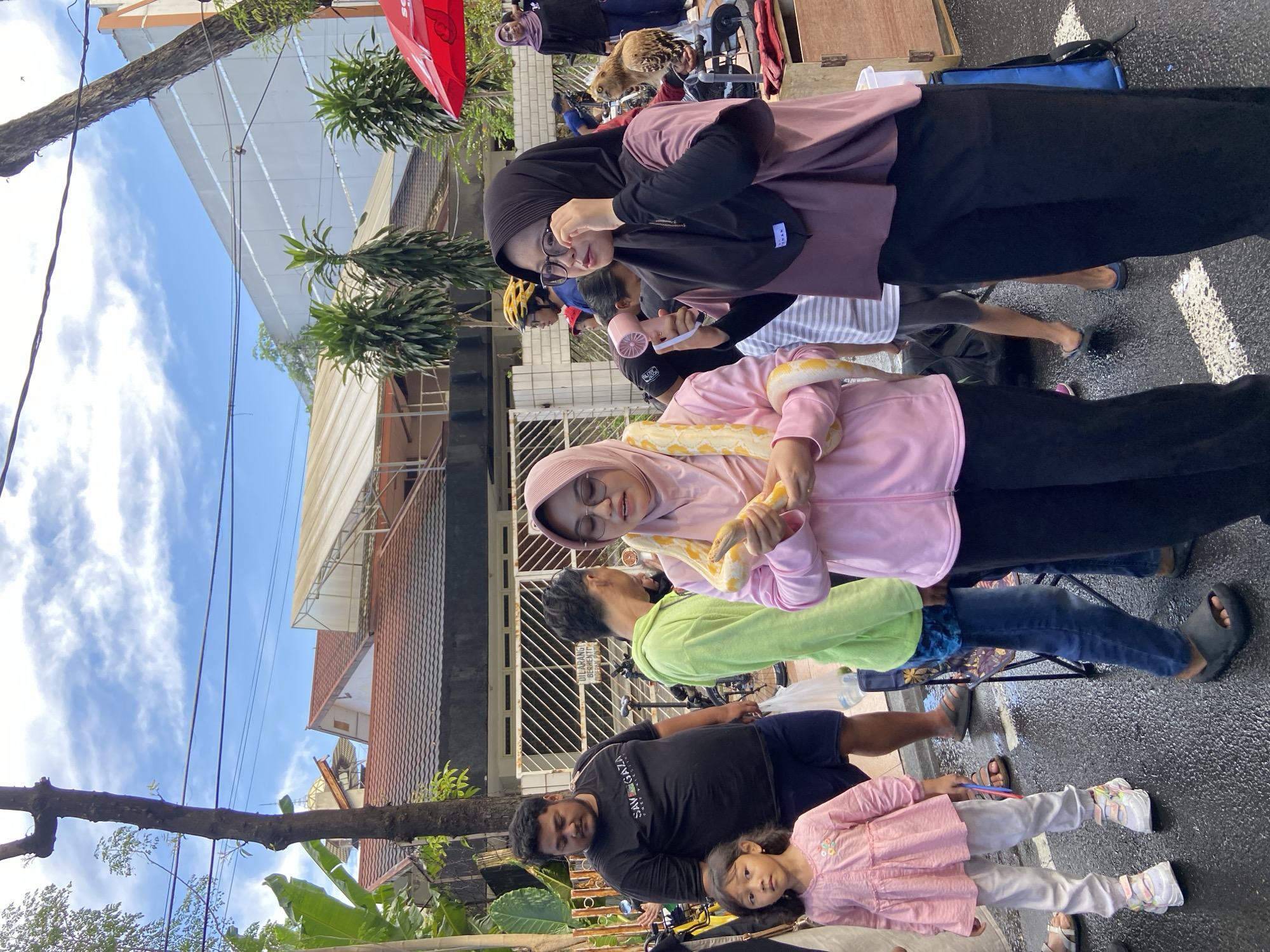 The image depicts a scene in Surabaya, Indonesia, with several individuals engaged in various activities. A woman wearing a pink hijab and matching top holds a yellow and white snake around her neck. Behind her stands a person in a green shirt. To the left, a man in a black shirt stands next to a young girl in a pink dress. Further back, a woman wearing a black hijab and glasses is looking at something. In the background, there are buildings with tiled roofs and a partially visible blue sky with some clouds. Street lamps and trees are also present in the background. The environment appears to be a street or public area.