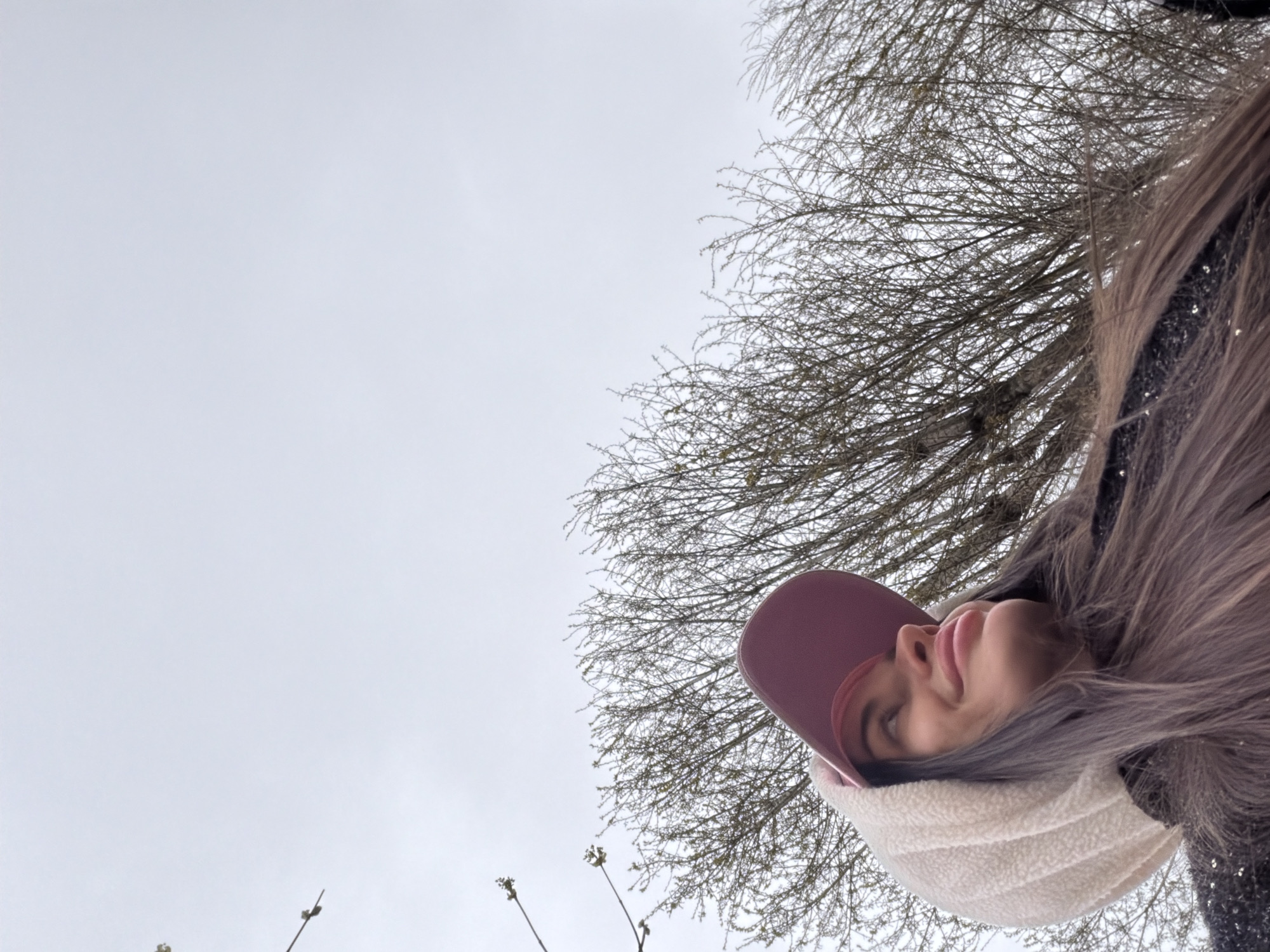 A woman is looking up into the camera, with trees behind her and a grey sky above. She is wearing a pink baseball cap and a cream-colored scarf. Her hair is brown and blonde. The location is Saint Petersburg, Russia.