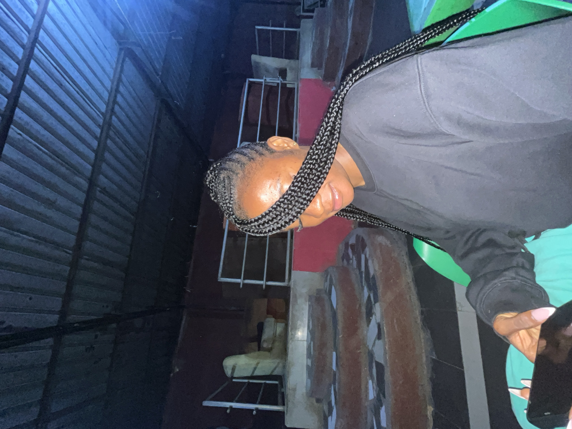 A young Black woman with braided hair that partially covers her face is seated outdoors at night, looking down at a black smartphone in her hands. She is wearing a dark crewneck sweatshirt. Her fingernails are painted a pale pink. She is seated in a bright green plastic chair. In the background, there is a dark corrugated metal roof supported by beams. Below the roof, there are steps with red and grey tiled surfaces, and some metal railings. The location is identified as Gura Top, Nigeria.