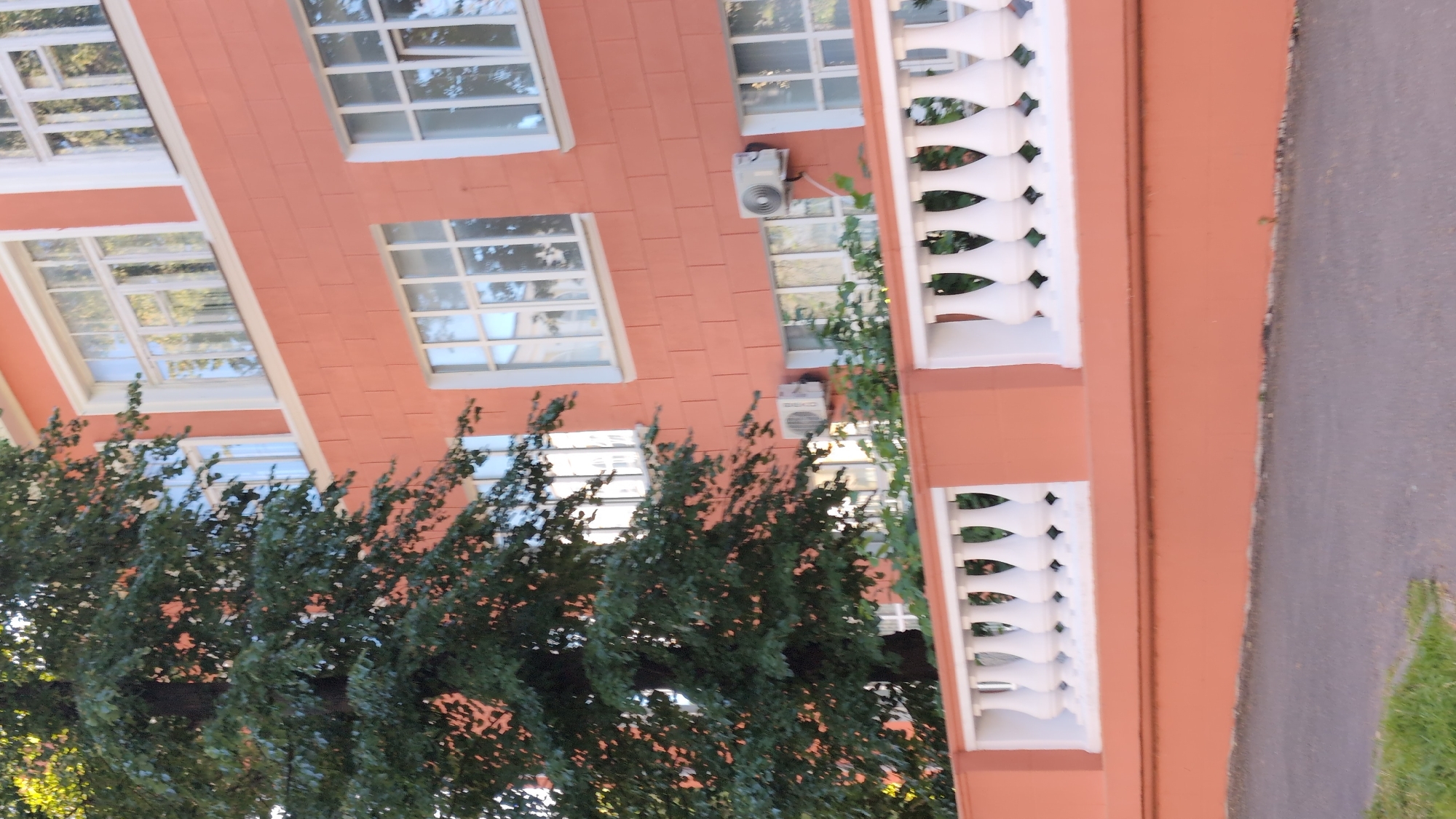 The image shows the exterior of a building with salmon-colored brickwork and white-trimmed windows. A white balustrade runs along the side of the building, which is partially obscured by green foliage from a tree. The building appears to have at least two stories, with rows of windows visible on each level. Two white air conditioning units are mounted on the exterior wall. The image is taken from an angle looking upwards at the building, with a paved surface visible to the right. The scene suggests an outdoor setting, possibly a street or courtyard. The lighting indicates daytime, and the presence of green foliage suggests it is not winter. The overall impression is one of a well-maintained building in an urban or semi-urban environment. No people are visible in the image. There is some faint text on the air conditioning units, but it is illegible. The location context provided is Nizhny Novgorod, Russia.
