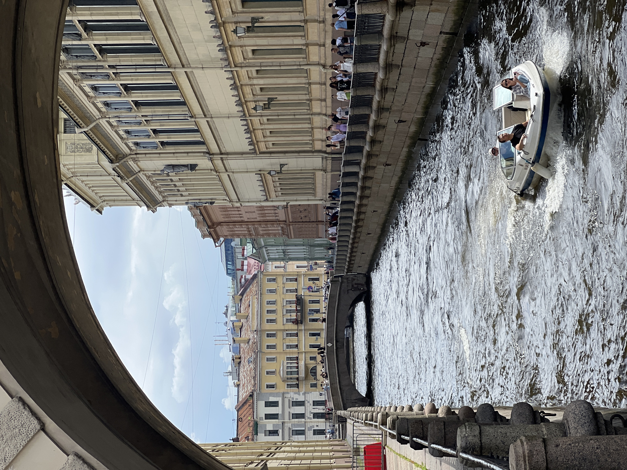 This image captures a scenic view of a canal in Saint Petersburg, Russia, likely the Griboyedov Canal, given its architectural style and the presence of a stone bridge and embankment with pedestrian traffic. A white motorboat with three occupants is cruising down the canal, creating a wake. The boat is moving from right to left in the frame.
On the right bank of the canal, a long, pale yellow building with classical architectural features lines the water. It has a series of large windows and decorative elements along its facade. A black metal railing runs along the top of the stone embankment. Numerous people are walking along the embankment, some in small groups and others individually.
On the left bank, a darker, multi-story building is visible, with a distinctively arched structure overhead, possibly a bridge or an overpass, partially obscuring the sky and distant buildings. The canal is flanked by stone embankments with decorative posts.
In the distance, a stone bridge with an arched span crosses the canal, with more buildings of varying colors and architectural styles visible beyond it. The sky is overcast with scattered clouds. The overall impression is a vibrant urban waterway scene with elements of historical architecture and leisurely activity.