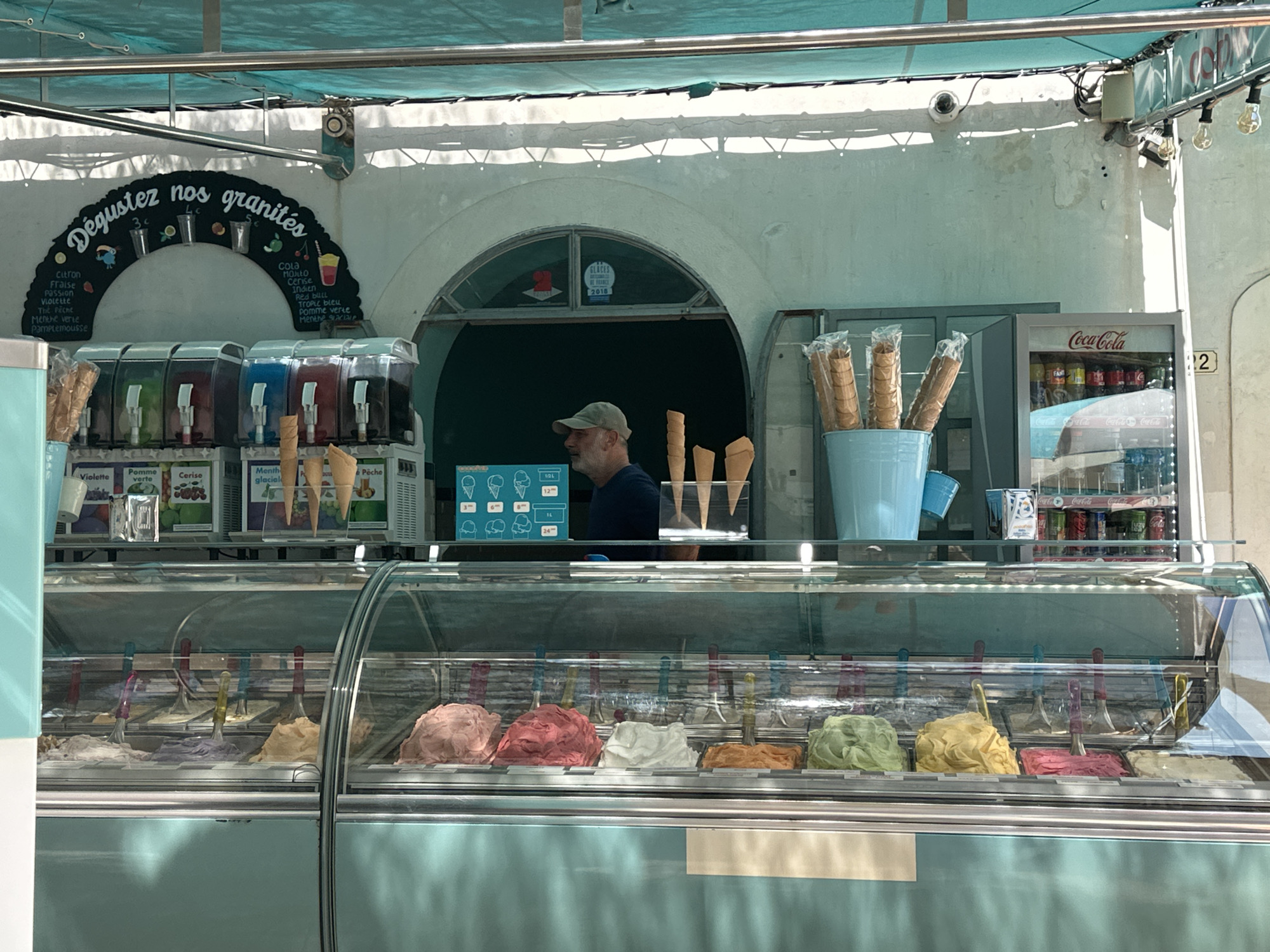 This is a medium shot of an outdoor ice cream stand in Hyères, France, possibly along the coast given the sunny ambiance. A man wearing a cap and a dark t-shirt is standing behind the counter. In front of him is a refrigerated display case filled with various flavors of gelato. To his left are several large, clear containers holding brightly colored frozen slushies. To his right, a blue bucket contains individually wrapped ice cream cones. Behind the man, a Coca-Cola refrigerator is visible, stocked with cans. Above the slushie machines, a black arched sign lists various flavors of "granités" (Italian ice). The overall environment suggests a warm, pleasant day, likely during tourist season. The architectural style of the building is simple, with an arched entryway. The lighting indicates it is daytime.