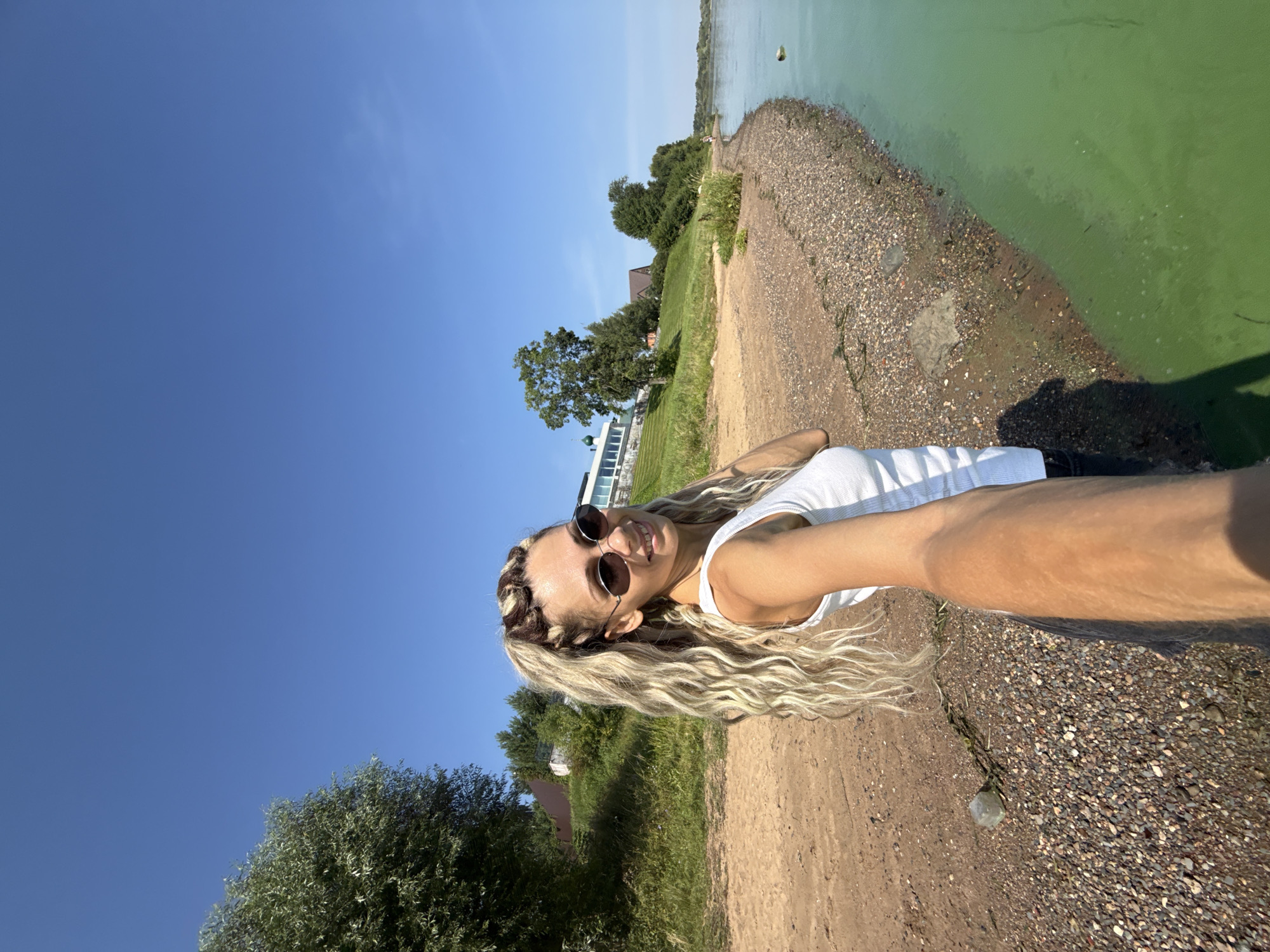 This is a selfie taken outdoors on a sunny day, likely in Rusanovo, Russia, given the context. A woman with blonde, long, wavy hair styled in a high ponytail and adorned with braids is in the foreground, extending her right arm towards the camera. She is wearing sunglasses and a white tank top. She is standing on a pebbly beach next to a body of water that appears green, possibly due to algae bloom. Behind her, there is a sandy area, a grassy incline with trees and buildings, and a clear blue sky. The woman is smiling and looking towards the camera, seemingly engaging with the viewer.