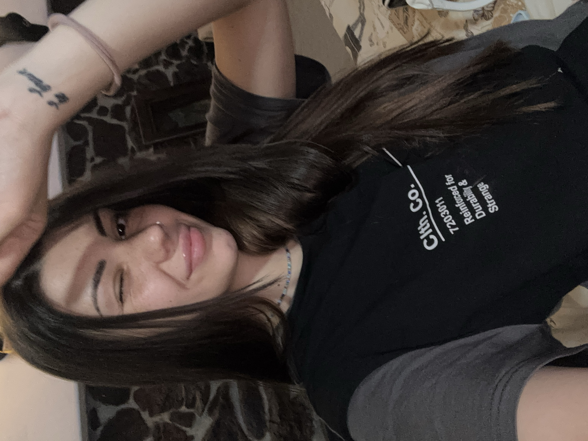A person with medium-length brown hair is lying down. The person is wearing a black t-shirt with white lettering that says "Clth. Co. 7203011 Reinforced for Durability & Strange." The person has a nose ring and is sticking out their tongue and winking. The person has a bracelet on their wrist and a tattoo on their forearm. The background appears to be a wall of dark, textured stone and patterned bedding. There is no information to suggest the location is Belopoptsi, Bulgaria.