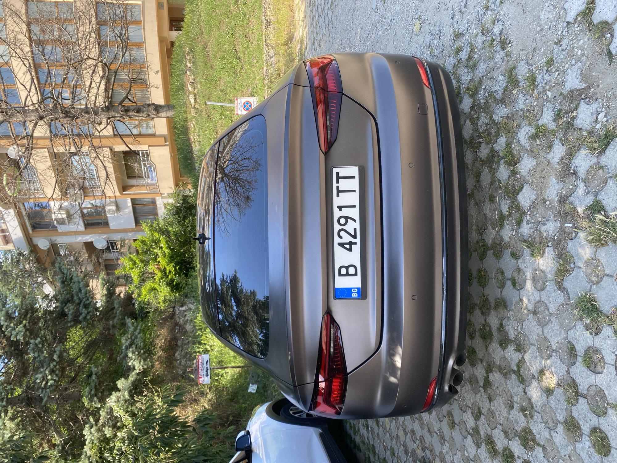 A matte brown Audi A5 is parked on a gravel and concrete interlocking block surface. The car has a Bulgarian license plate that reads "B 4291 TT". In the background, there is a multi-story apartment building with balconies and windows. Adjacent to the building is a grassy slope with trees. To the left of the car, a portion of a white SUV is visible. There are two red and white warning signs planted in the ground, one on the left side of the image and another on the right, both featuring Bulgarian text and symbols indicating parking restrictions. The lighting suggests it is daytime.