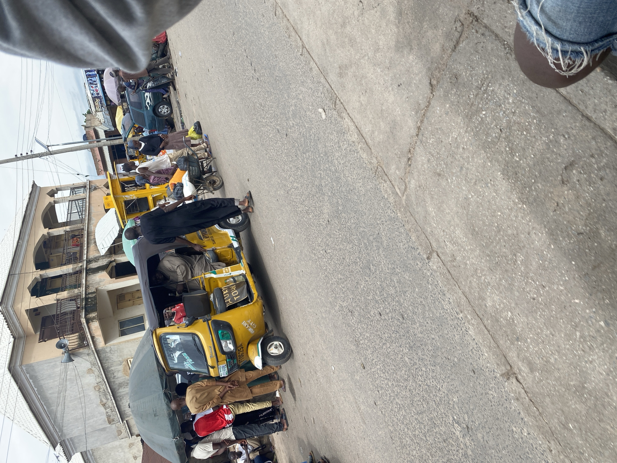 A yellow motorized tricycle, commonly known as an "okada" or "keke," is parked on a paved street. Several people are around the tricycle, some appearing to be passengers and others possibly the driver or vendors. One person in the foreground is dressed in black, partially obscuring the tricycle's front. Another group of people are seated on the side of the tricycle, while others stand nearby. The tricycle has some visible text on its side, including "ONITSHA" and what appears to be a license plate with letters and numbers. The building behind the tricycle is a multi-story structure with balconies and windows. The scene suggests a typical street market or transportation hub. The lighting indicates it is daytime. The overall atmosphere appears to be busy and active, typical of a commercial area in Jalingo, Nigeria.