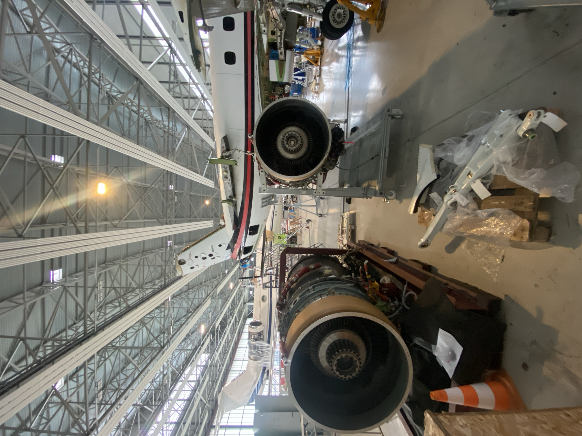 The image depicts an aircraft maintenance hangar. The location is identifiable as Skulte, Latvia due to the type of aircraft. The interior of the hangar is characterized by a high ceiling with exposed metal framework and overhead lighting. The primary subject is an aircraft with one engine mounted and another removed. The removed engine is present and appears to be undergoing maintenance. Various tools, scaffolding, and disassembled parts are scattered around the hangar floor. No people are visible in the image.