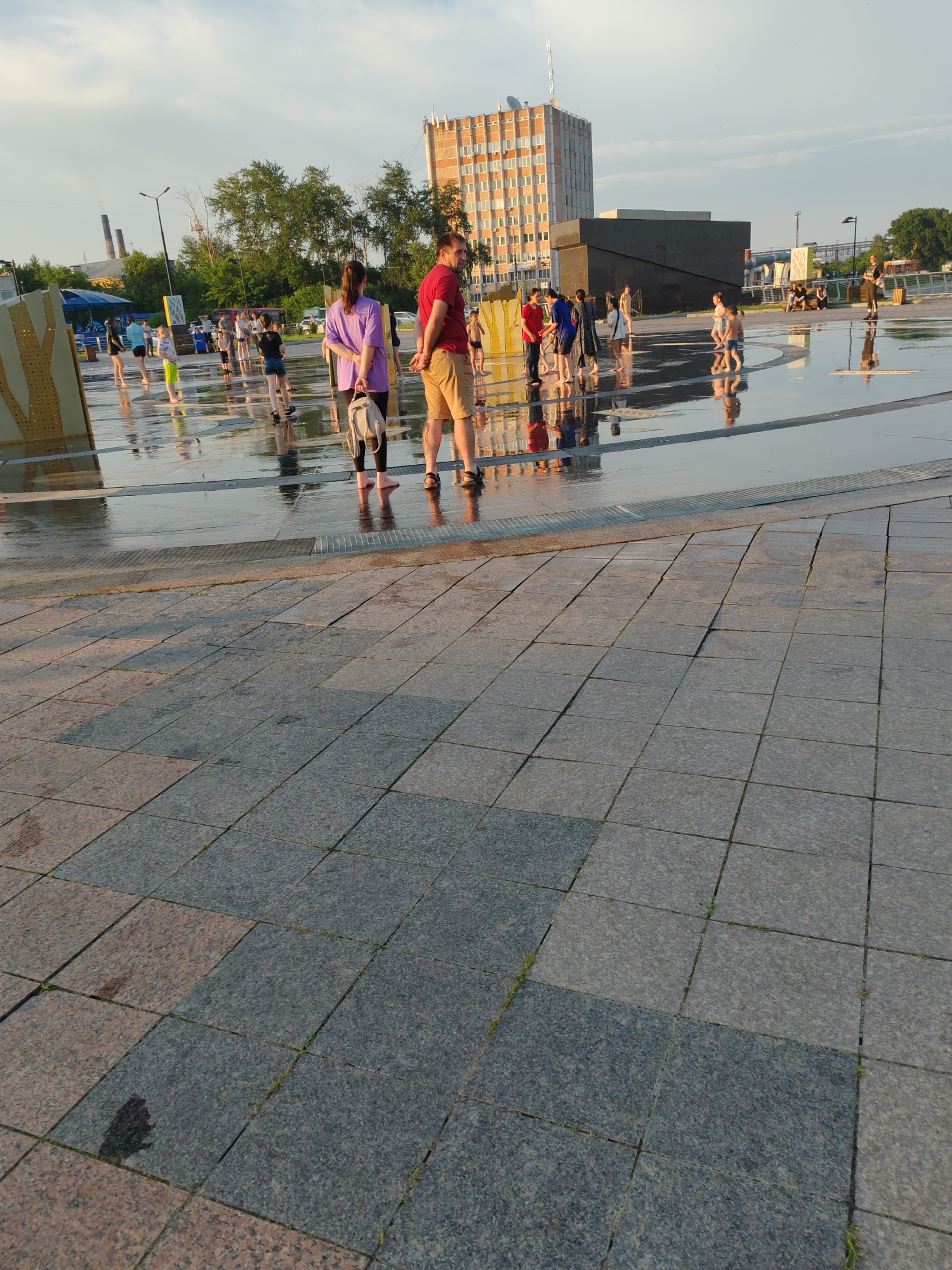 In Komsomolsk-on-Amur, Russia, a group of people are enjoying a public fountain area on a sunny day. The foreground is paved with large, grey granite tiles. In the mid-ground, a wide, shallow fountain area is covered in a thin layer of water, reflecting the sky and the surrounding environment. Several people are standing in the water, some appearing to be children playing, while others are adults observing or walking through the shallow water.
A man in a red t-shirt and beige shorts stands facing to the right, with his hands behind his back. To his left, a woman in a purple t-shirt and dark leggings holds a light-colored backpack. Further into the fountain area, other individuals are seen, including children in colorful clothing and adults in casual attire. In the background, a tall, multi-story building with a distinctive brick facade stands prominently, alongside other urban structures and trees. The sky above is partly cloudy, with the warm light of the sun casting reflections on the wet surfaces.