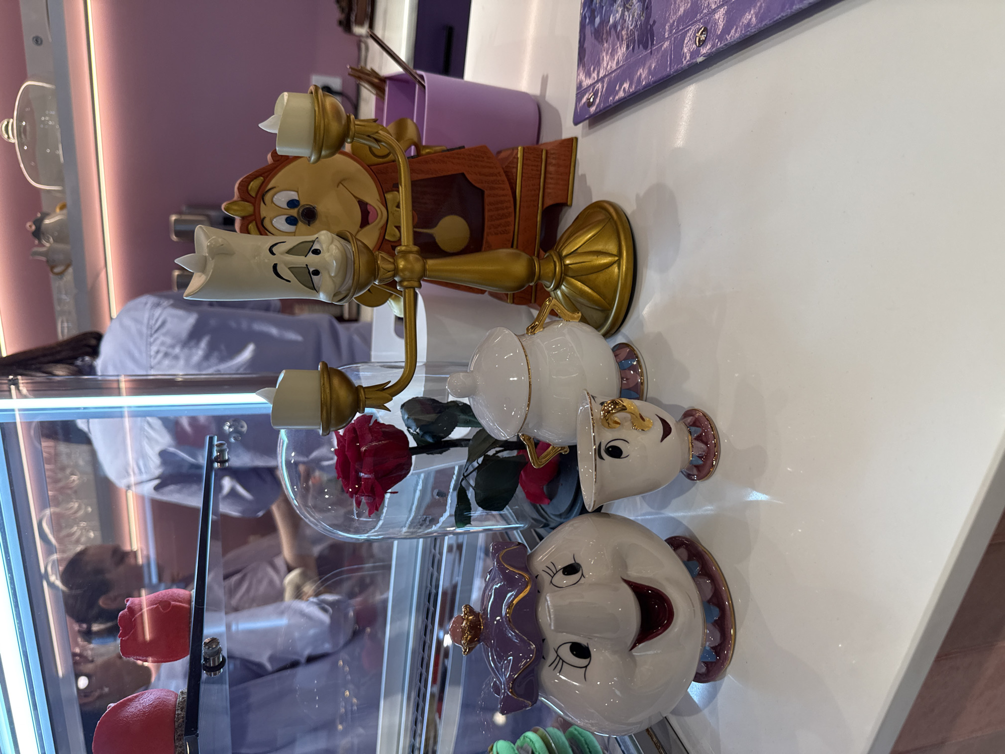 The image shows a display featuring "Beauty and the Beast" character figurines within a glass case. The characters include Cogsworth (the clock), Lumière (the candlestick), Mrs. Potts (the teapot), and Chip (the teacup). There are also representations of the enchanted rose in a glass bell jar. The display appears to be in a retail or bakery setting, as suggested by the macarons visible near the bottom of the glass case and the person wearing light-colored clothing in the background. There is no evidence of it being located in Vladivostok, Russia.