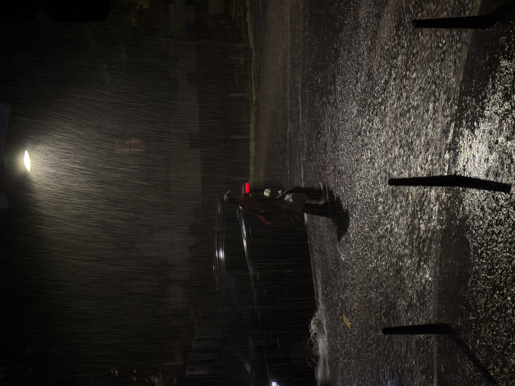 A person on a scooter rides through a flooded street at night in Belgrade, Serbia, under a street lamp during heavy rainfall. The water is ankle-deep in some areas, with ripples and splashes visible. The person, seen from behind, is wearing a dark jacket and carrying a bag over their shoulder. A dark-colored car is parked on the side of the street, its taillights faintly visible. The scene is dimly lit by the street lamp, casting a warm glow on the rain and creating a moody, atmospheric effect. The background is dark and indistinct, with the suggestion of buildings and trees.
