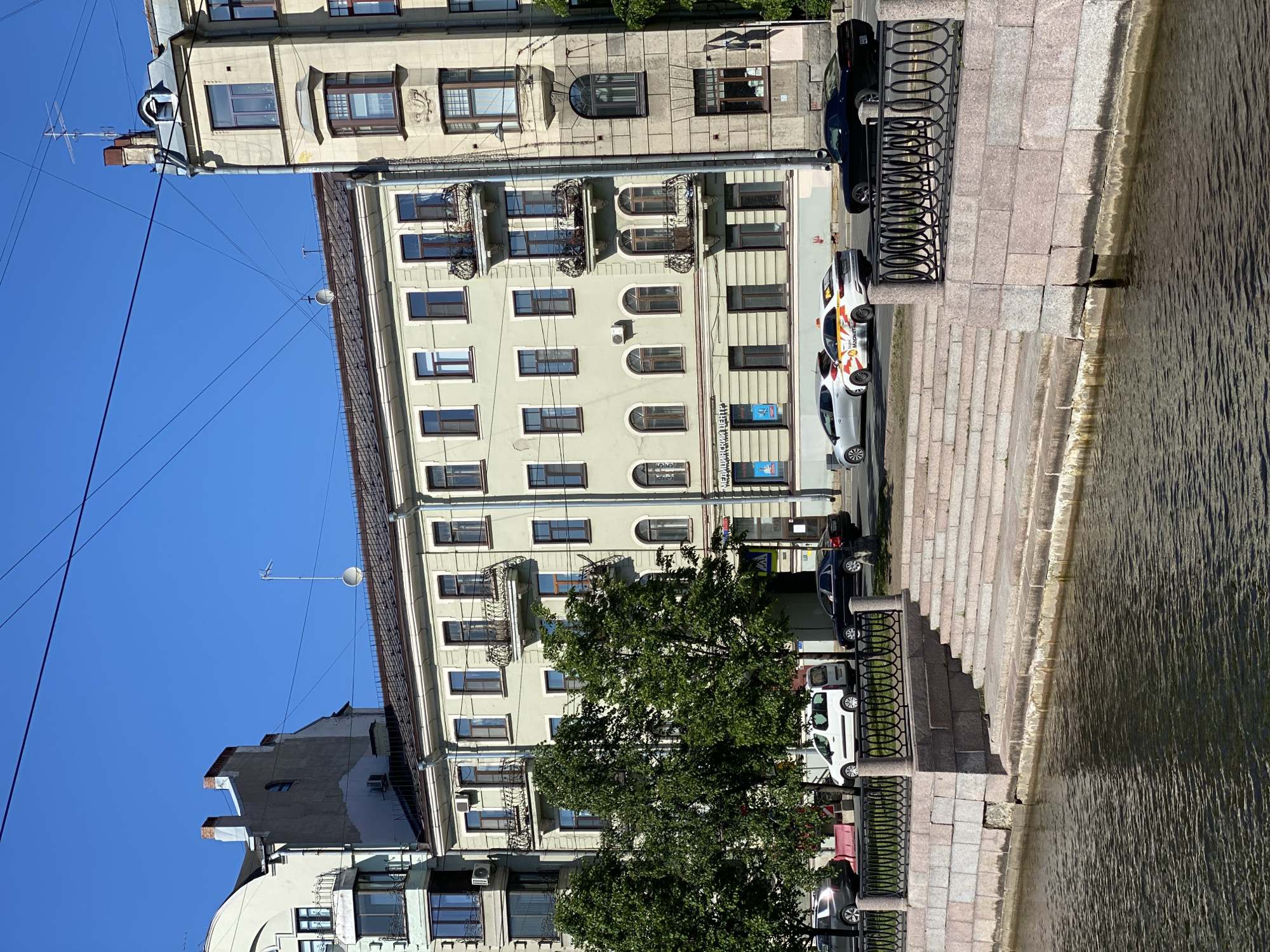 This vertically oriented image captures a bright, sunny urban scene alongside a canal, likely in Saint Petersburg, Russia.
In the foreground, on the right, is a calm canal or river with ripple patterns on its surface, reflecting the sunlight. An ornate black metal railing separates the water from a wide stone embankment with several steps leading down to the water.
Moving left from the embankment, there's a paved road or street. Several cars are parked along the curb. Notable among them are two white cars with yellow and orange branding, clearly displaying the word "Яндекс" (Yandex) and likely "Такси" (Taxi), indicating they are Yandex Taxi vehicles. A dark sedan and a white utility van are also visible. A large, leafy green tree partially obscures the view of the buildings behind it.
The background, on the left side of the image, features a row of multi-story buildings, typical of historical European architecture. The buildings have pale, light-colored facades (some cream, some light green/grey), numerous windows, and decorative balconies with wrought-iron railings. Some windows are arched, while others are rectangular. A drainpipe runs vertically down one building. Overhead, against a clear, bright blue sky, numerous utility wires crisscross, and an antenna is visible on the rooftop of one building.
The scene depicts a clear, sunny day, likely morning or early afternoon, given the bright light and distinct shadows. No people are visible in the frame, giving the scene a quiet, serene atmosphere. On a building facade, a blue sign with Cyrillic text, possibly "РЕМОНТ" (Repair) or "АРЕНДА" (Rent), is discernible, along with other small, unreadable signs.